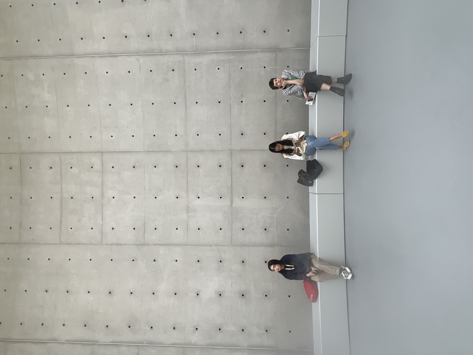Three women are seated on a light gray bench in front of a large, light gray concrete wall with visible circular indentations and horizontal seams. The woman on the left is wearing a dark blue hooded sweatshirt and tan pants, with a red bag to her left. The woman in the middle is wearing a white top and blue jeans, holding a book. The woman on the right is wearing a black and white striped shirt and black pants, also holding a book and resting her chin on her hand. A black bag is placed between the woman on the left and the woman in the middle, and another black bag is to the right of the woman in the middle. The floor in front of the bench is a smooth, light gray surface. The overall setting appears to be modern and minimalist. The image was taken in Paris, France, specifically at the Institut du Monde Arabe.