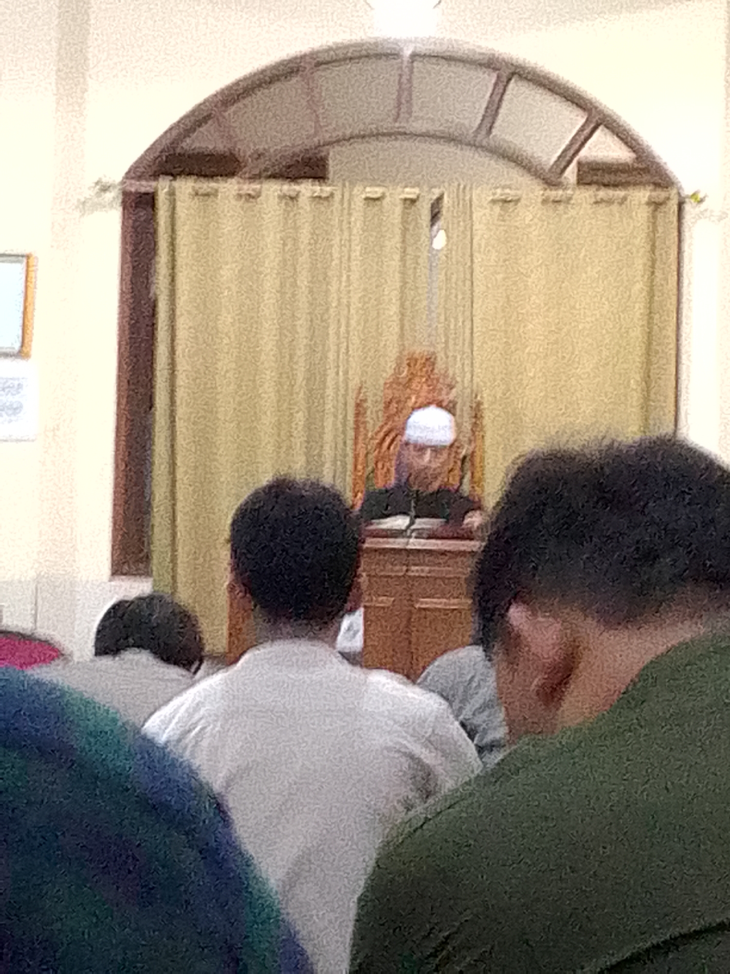 The image depicts a group of people gathered in what appears to be a mosque in Kertajaya, Indonesia. In the background, a man wearing a white cap and a dark robe is seated behind a wooden podium, likely delivering a sermon or leading prayers. He is facing a book placed on the podium. The man is positioned behind a light brown curtain. In the foreground, several individuals are visible from behind, facing the man in the background. One person in the center is wearing a white shirt, another on the left is wearing a dark top, and a person on the far right is wearing a dark green garment. The environment appears to be indoors, with arched window-like structures visible above the curtains. The lighting is soft, suggesting an interior space.