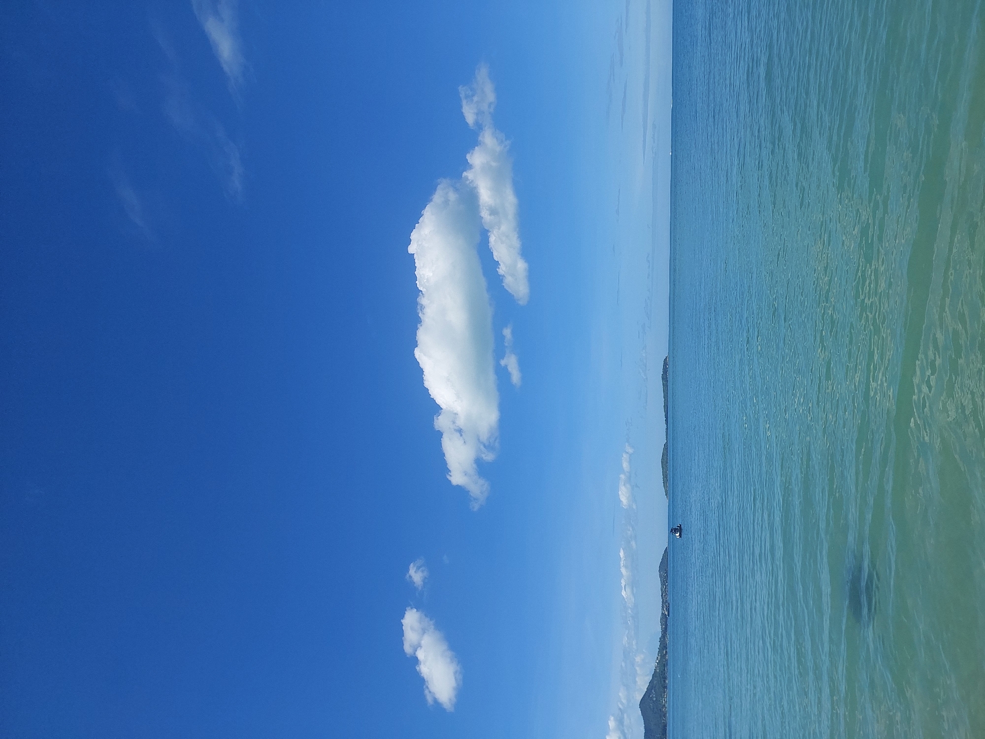 This is a landscape photograph of a bright, clear day at Baan Chaweng Noi, Thailand. The image is dominated by a vast expanse of vivid blue sky with scattered, fluffy white clouds. The foreground is comprised of calm, turquoise ocean water, reflecting the sunlight with gentle ripples on the surface. In the distance, the coastline is visible with a lush green, mountainous island on the left. Several smaller islands and a strip of land with buildings can be seen along the horizon. A small boat is positioned in the middle of the water, appearing to be moving. The overall impression is one of tranquility and natural beauty.