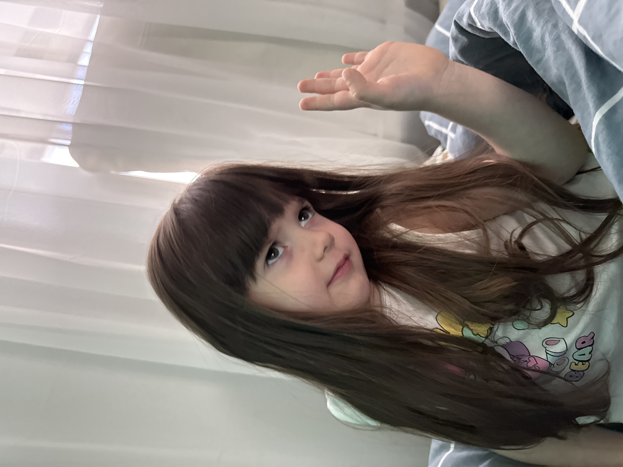 A young girl with long, dark brown hair is lying in bed, partially covered by a grey and white striped blanket. She is looking up and to her left, with her right hand raised slightly. Her expression appears thoughtful or curious. The background consists of sheer, white curtains that let in soft light, suggesting it might be daytime. The girl is wearing a white t-shirt with colorful cartoon characters and text that reads "BEAR". The scene is intimate and relaxed, conveying a sense of comfort and perhaps a moment of playfulness or contemplation.