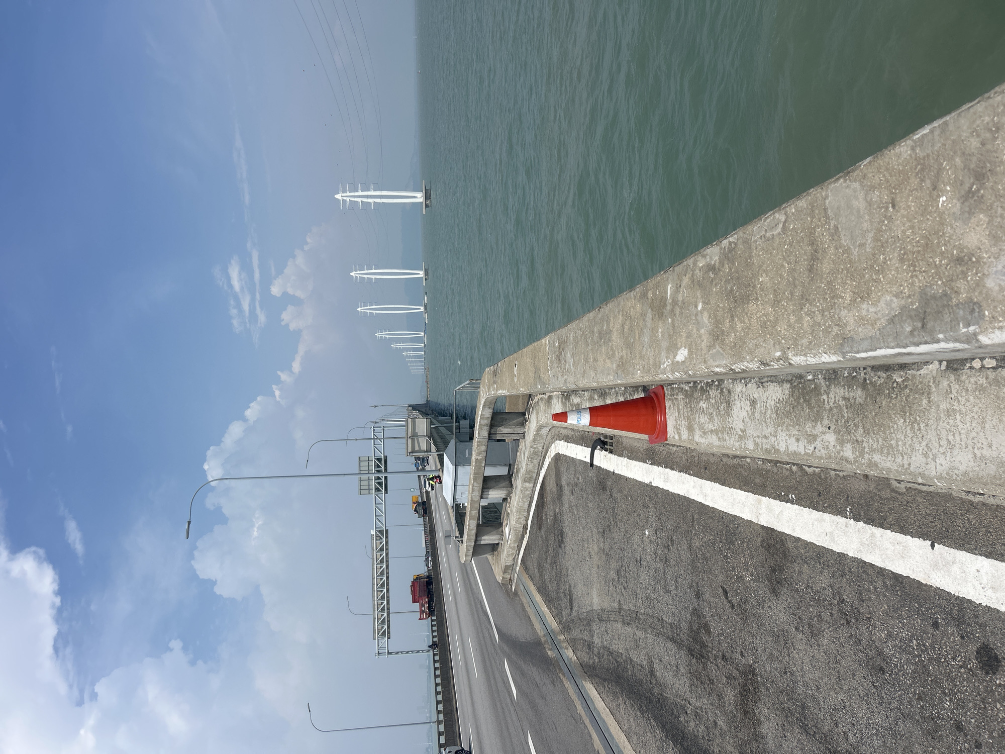 The image captures a view of a section of the Penang Bridge, connecting George Town, Malaysia, to the mainland. The bridge is an elevated structure over a body of water, appearing to be the sea. On the right side of the image, a concrete barrier runs along the edge of the bridge, overlooking the greenish-blue water. A single orange traffic cone with the word "POLIS" printed on it is placed near the barrier, suggesting a temporary traffic control or a safety measure.
The roadway of the bridge is visible on the left, with lane markings and tire marks indicating vehicular traffic. Several vehicles are present on the bridge, including a red truck and other cars, some appearing to be moving. Overhead, streetlights and traffic signage are installed along the bridge. In the distance, a series of white, arch-shaped pylons supporting power lines stretch across the water, marking the continuation of the bridge or a nearby infrastructure. The sky is partly cloudy with a bright blue expanse. The overall scene depicts a sunny day on a major transportation artery in George Town, Malaysia.
