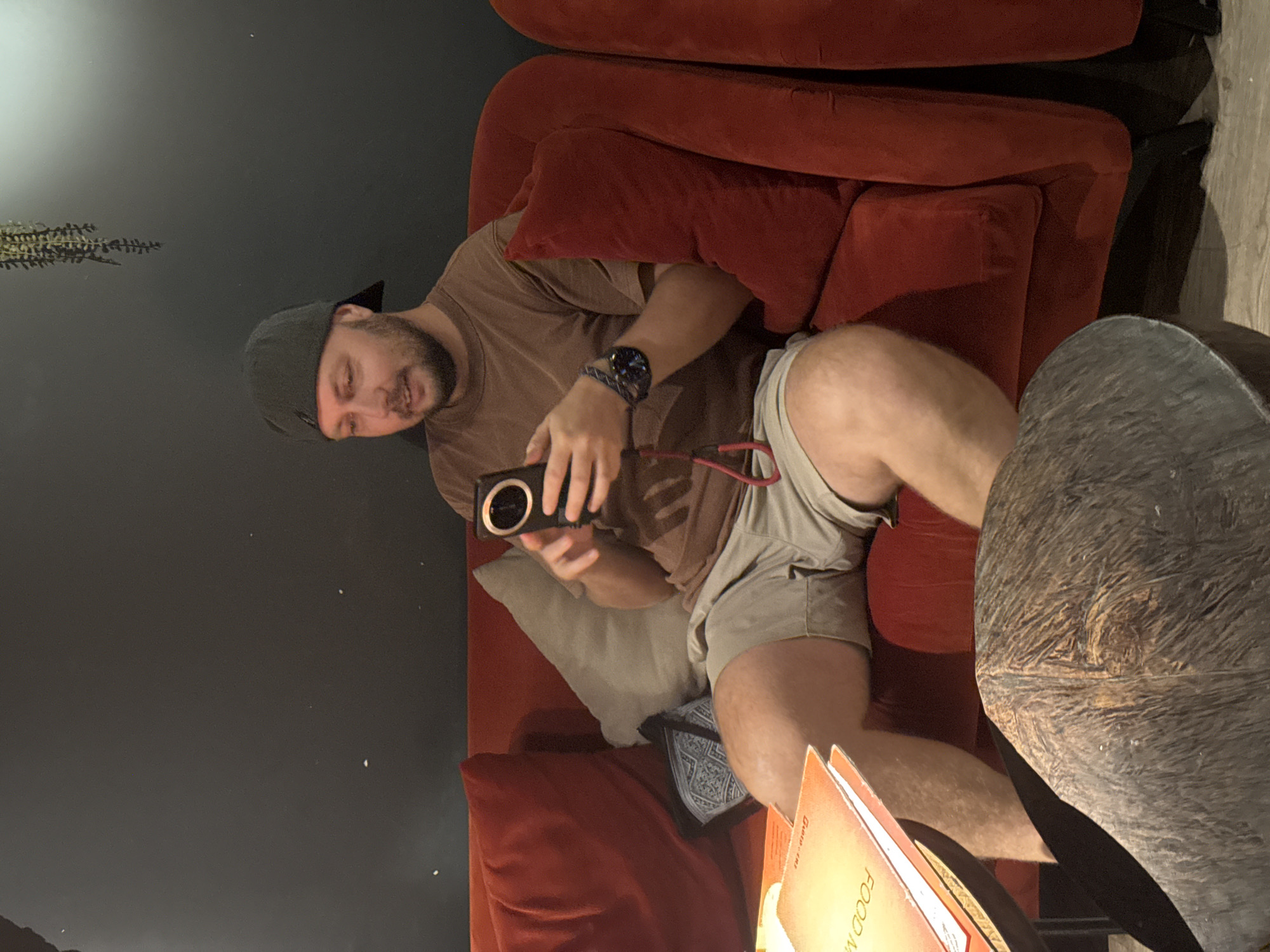 A man wearing a brown t-shirt, light-colored shorts, and a grey baseball cap is sitting on a red sofa. He is holding a black camera with a red strap attached. The man has a beard and appears to be looking down at the camera. A grey pillow is placed behind him on the sofa. To the right of the man, part of a dark, textured surface is visible, possibly a table or ottoman. In the foreground, several orange menu-like items are stacked, with some text visible, including "FOOD M". The background is dark, with a hint of a decorative plant on the left side. The lighting suggests an indoor setting, possibly a restaurant or lounge, with dim ambient light and some brighter spotlights. The overall mood appears relaxed and casual. The orientation of the image is portrait, with the top of the frame on the left. The presence of menus and a comfortable seating area suggests this could be a dining or leisure establishment in Hà Nội, Vietnam.