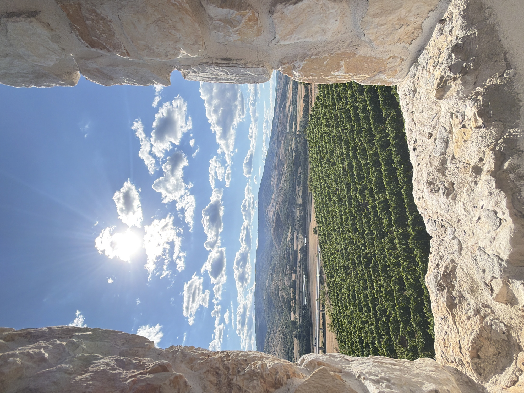 The image shows a view through a stone archway, looking out over a vineyard. The sky is blue with scattered white clouds and the sun is shining brightly. In the distance, rolling hills are visible, with a highway bridge crossing a small river in the middle ground. The foreground is dominated by rough stone walls, framing the scenic vista. The location is identified as Kocherinovo, Bulgaria. There are no people or animals present in the image. The scene is a landscape view of agricultural land and natural scenery, viewed from a historical structure.