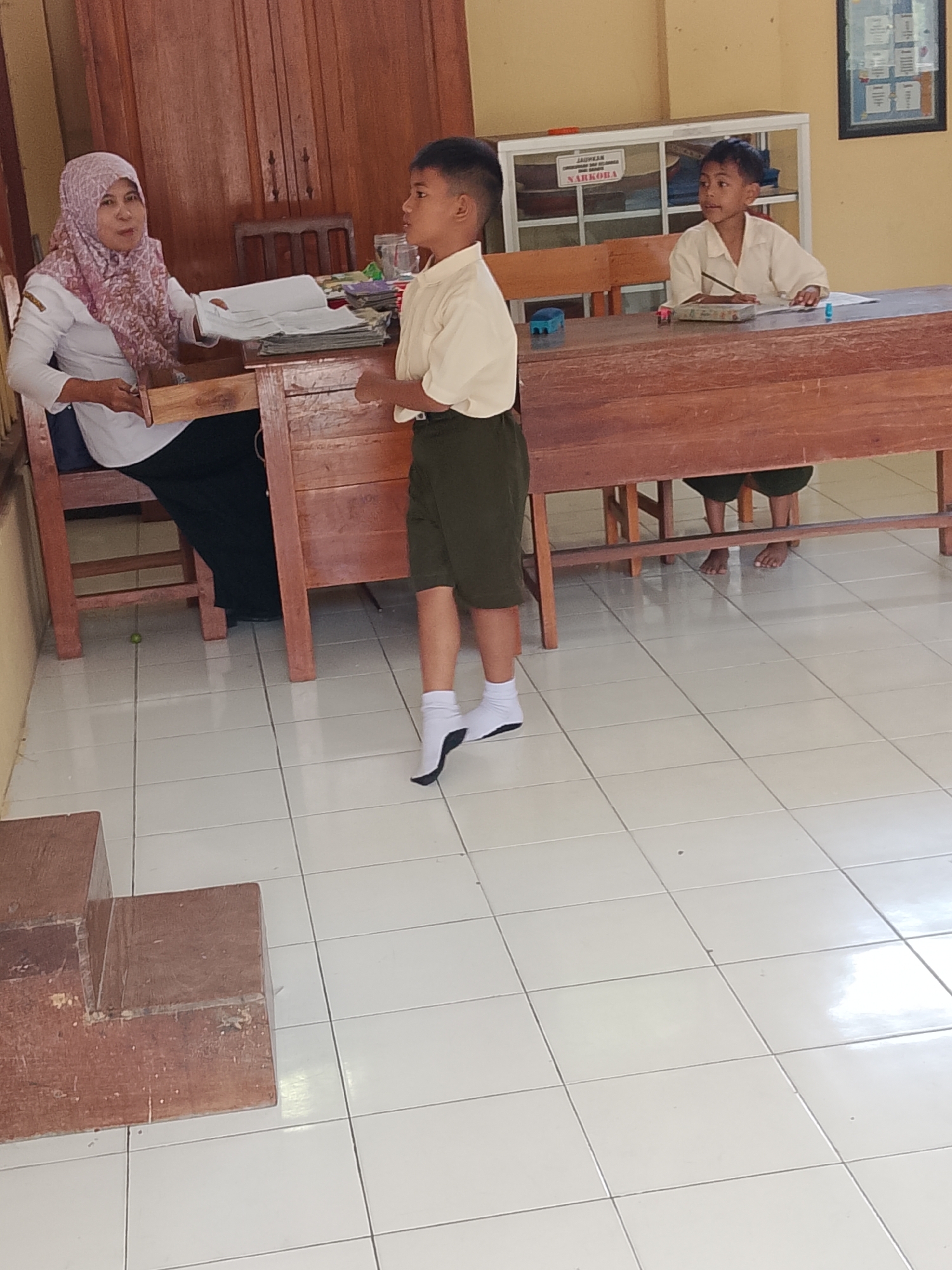 The image depicts a classroom or office setting. In the foreground, a young boy in a light-colored shirt and dark shorts stands facing left, with white socks and black shoes. To his left, a woman wearing a pink hijab and white shirt sits behind a wooden desk, holding papers. To the right of the standing boy, another young boy in a similar light-colored shirt sits at a desk with a pencil. The environment includes wooden furniture, tiled flooring, and a wall with a framed notice. The scene appears to be within a school or public building in Kepuh, Indonesia, given the context provided.