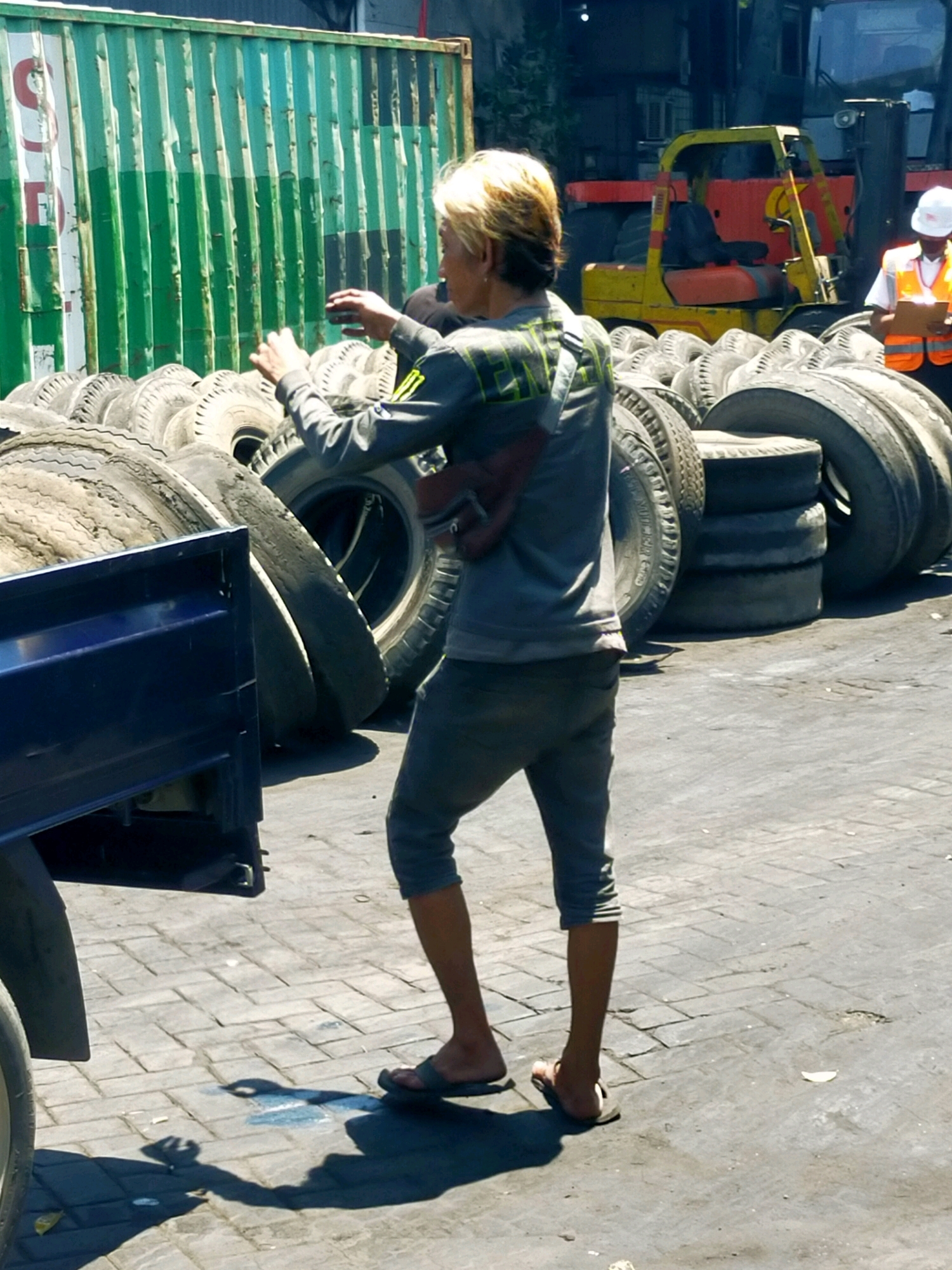 The image depicts an outdoor scene in Surabaya, Indonesia, likely a junkyard or storage area for used tires. In the foreground, a person with blonde hair is captured from the waist down, wearing a gray long-sleeved shirt and dark gray capri pants. They are barefoot and appear to be in motion, with their hands raised and extended forward. Their stance and posture suggest they are either reaching for something or engaged in some form of activity. Behind this individual, a large pile of used tires is visible, some stacked horizontally and others vertically. A blue truck bed is partially visible on the left side of the frame, also containing tires. In the background, a forklift with a yellow frame and red accents is parked, with a person seated in the driver's seat wearing a blue shirt. To the right of the forklift, another person is standing, wearing a white helmet and an orange vest, holding a clipboard. The ground is paved with cobblestones or similar interlocking stones. A large green shipping container is prominent on the far left, with vertical lines suggesting its corrugated structure. The lighting suggests it is daytime, with strong shadows cast by the objects and people. The overall impression is one of an industrial or commercial area where salvaged materials are handled.