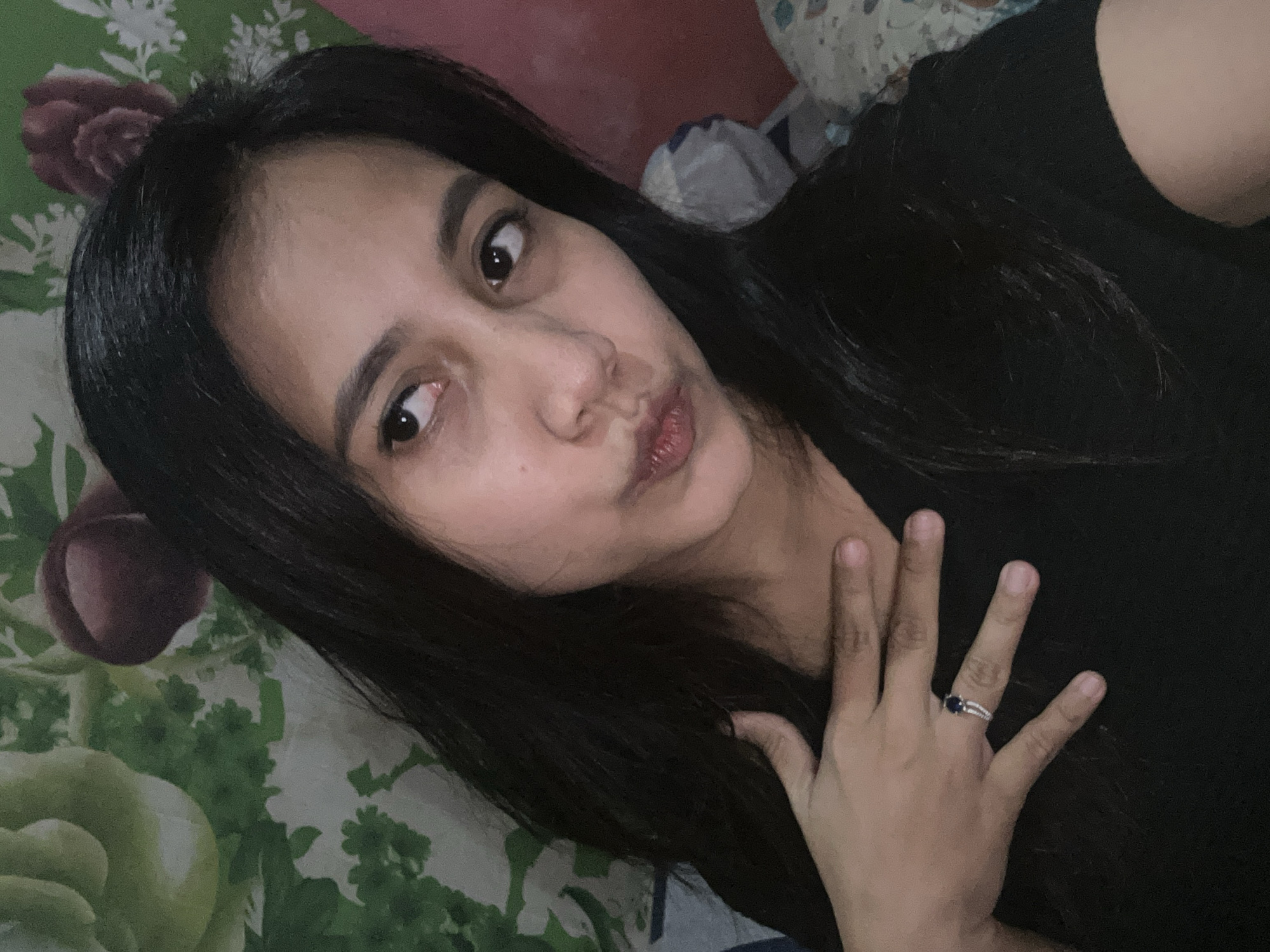 A young woman with long, dark hair and fair skin is shown in a close-up, selfie-style shot. She is wearing a black t-shirt and a ring with a dark blue stone on her left hand, which is placed on her chest. Her head is tilted to the right, and she is looking away from the camera with a slightly pursed expression. The background appears to be a bed with patterned bedding and pillows. The room has a floral wallpaper. The scene suggests an indoor setting, possibly a bedroom, in Depok, Indonesia.