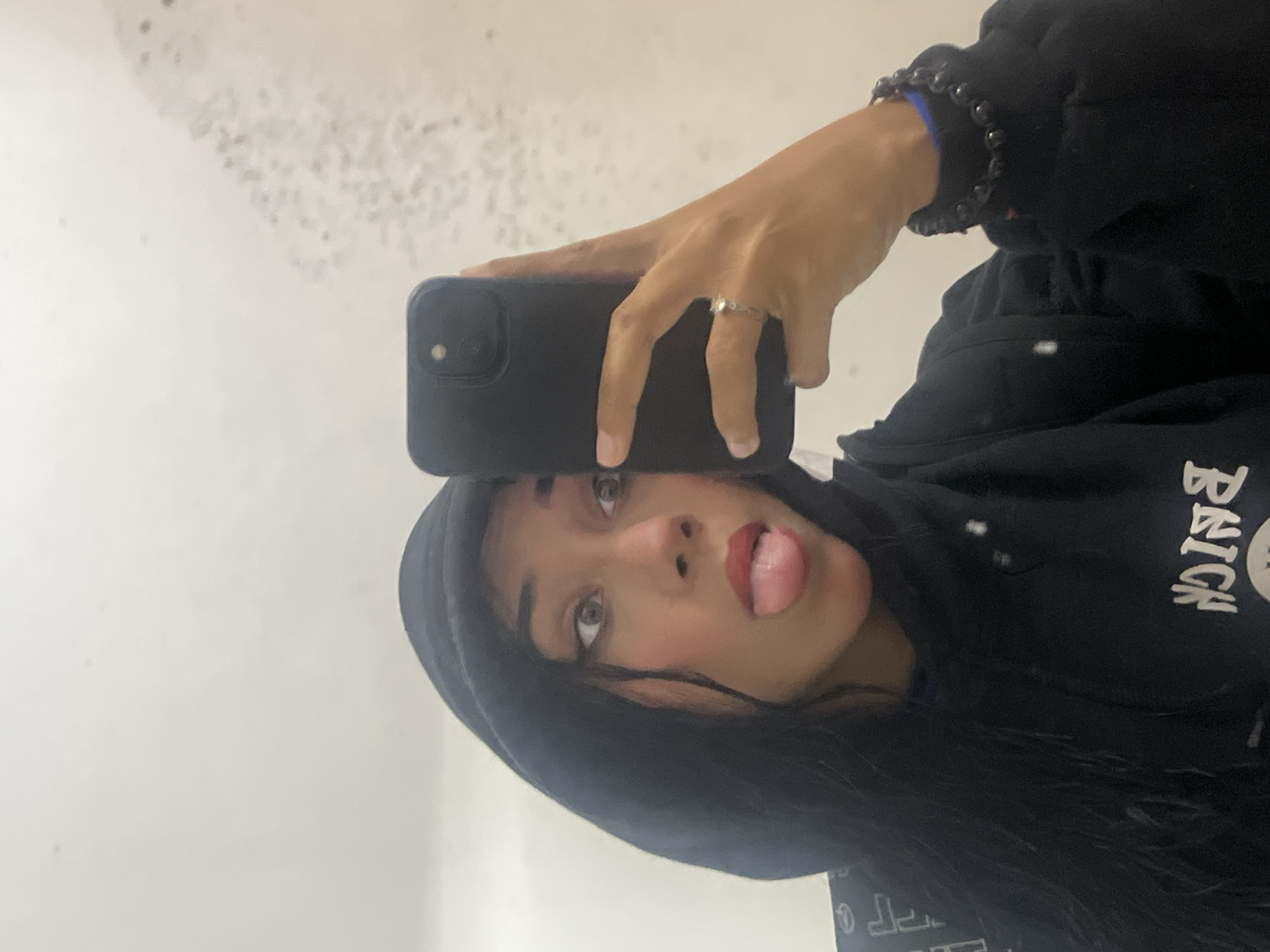 A person is taking a selfie using a black phone with two camera lenses. The person has fair skin, dark hair, and is wearing a black hooded sweatshirt. A black beaded bracelet is visible on the person's left wrist. The person is sticking their tongue out and looking at the camera. The background appears to be a white wall. The exact location of the image in Pampatar, Venezuela cannot be determined from the image.