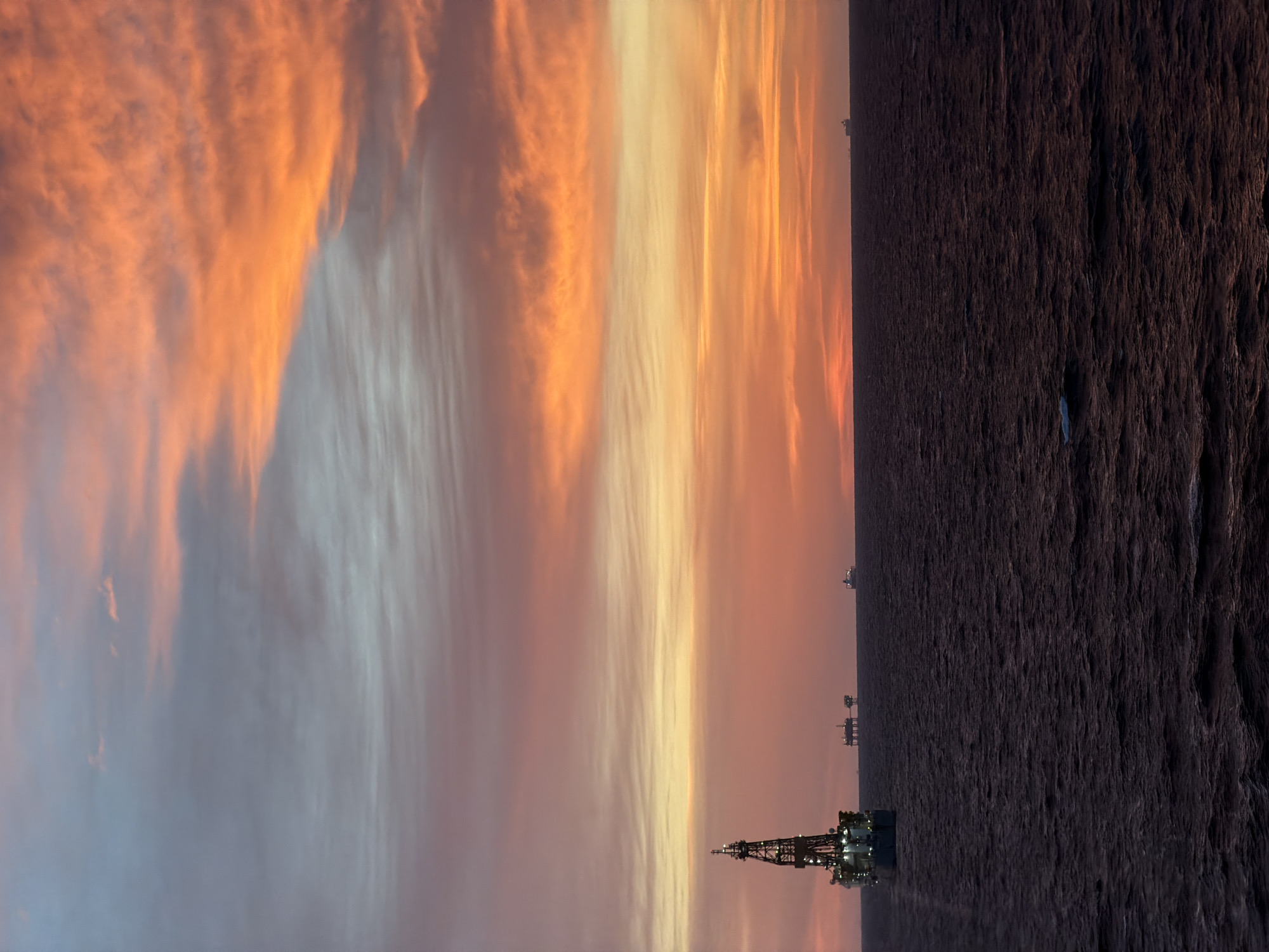 The image displays an expansive offshore industrial landscape captured during either sunrise or sunset. The upper third of the frame is dominated by a dramatic sky filled with long, wispy clouds, painted in striking hues of orange, yellow, pink, and soft blue, suggesting the sun's low angle.
Below the colorful sky, the vast expanse of the ocean occupies the majority of the image. The water surface appears dark and textured, with visible ripples and waves creating a rugged appearance.
Several offshore oil rigs or drilling platforms punctuate the seascape. A prominent, brightly lit drilling platform stands in the left foreground, its intricate structure detailed against the vibrant sky. Further in the mid-distance, multiple smaller platforms are scattered across the water, also illuminated by artificial lights, indicating ongoing operations. A small, indistinct light-colored object floats on the water in the right mid-ground.
The scene combines the natural beauty of a dawn or dusk sky with the stark reality of industrial activity on the open sea. The lighting on the rigs contrasts with the fading or emerging natural light. No specific human activity or figures are discernible, and no text is visible.