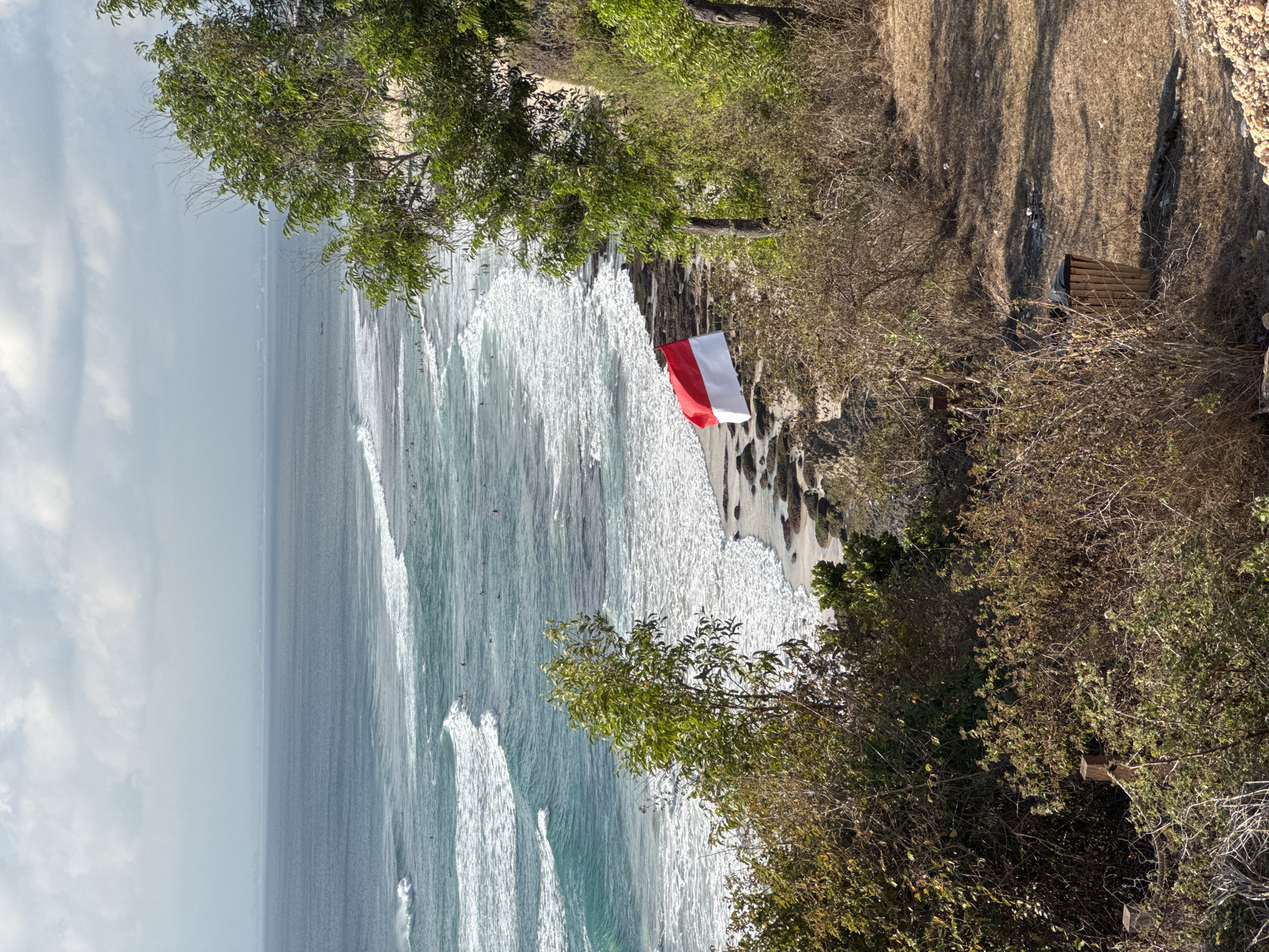 The image depicts a scenic coastal view in Kuta Selatan, Indonesia, with a focus on a stretch of ocean and a rocky shoreline. The water is a vibrant turquoise, with white-capped waves rolling towards the shore. Several surfers are visible in the water, scattered across the waves.
On the rocky shore, a red and white flag, the flag of Indonesia, is prominently displayed, appearing to be attached to a makeshift pole. The flag is positioned on a slightly elevated rocky area overlooking the ocean.
The foreground and right side of the image are dominated by lush green vegetation and dry brush, suggesting a natural, somewhat untamed environment. A rustic trash bin made of bamboo is visible in the lower right quadrant, adding a touch of human presence to the otherwise natural setting. The sky above is partially cloudy, with diffused sunlight indicating a bright but not intensely sunny day. The overall scene conveys a sense of a popular surfing spot and a patriotic display by the Indonesian flag.