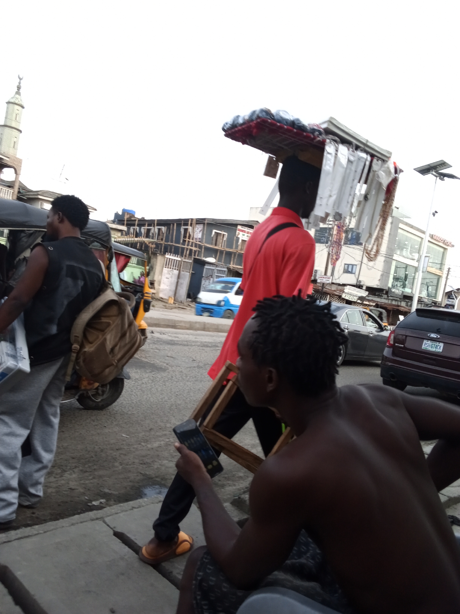 The image captures a vibrant street scene in Ado, Nigeria, depicting the daily life and commerce of the area. In the foreground, a man with dreadlocks, shirtless and wearing patterned shorts, is seated on a low surface, engrossed in his mobile phone. His back and right shoulder are visible, showing a muscular physique.
To his left, a man in a bright red shirt walks towards the viewer, balancing a large, loaded tray of merchandise on his head. The tray appears to hold various small items, possibly phone accessories or small electronics, indicative of a street hawker. He wears orange flip-flops.
Further to the left, another man, dressed in a black sleeveless top and grey trousers, stands by a yellow and black Keke Napep (tricycle taxi). He carries a brown backpack and appears to be either waiting or moving along the street.
The background reveals a bustling urban environment. A distinctive mosque minaret with a crescent moon at its peak rises on the far left. Various buildings, some appearing residential and others commercial, line the street. One building shows signs of construction with visible scaffolding. The road is busy with traffic, including a dark sedan and a maroon SUV. The SUV's license plate, "PSH 67ACK", is clearly visible. Other faint text on buildings includes what might be "LABORATORY" or "TYCOM" and "OYALEK".
The sky is bright and overcast, suggesting daytime. The overall impression is one of a typical, busy day on a Nigerian street, filled with movement, street vendors, and local transport.