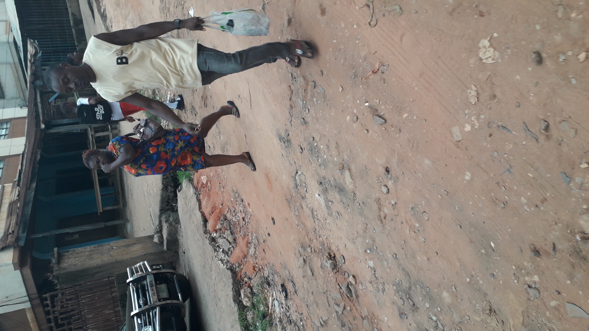 The image shows two individuals walking on a partially paved, dirt road in Obosi, Nigeria. One individual, a man, is wearing a light-colored t-shirt, jeans, and sandals. He is carrying a plastic bag. The other individual, a woman, is wearing a floral dress and flats. The road is cracked and uneven, with debris scattered on the surface. Buildings are visible in the background, suggesting an urban or semi-urban setting. A vehicle is parked on the side of the road.