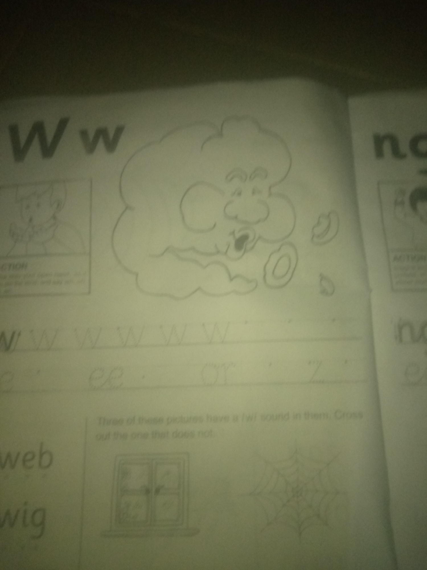 The image is a close-up, angled shot of a page from what appears to be an alphabet workbook. The page features the letter "W" prominently displayed in large print at the top left. To the right of the letter "W" is a cartoon drawing of a cloud-like figure with a face, appearing to be blowing outwards, with several circular shapes (possibly cookies or doughnuts) scattered around it. Below the large "W" are lines with handwritten cursive letters, including "W" and other cursive characters that are difficult to discern due to image quality. Further down, the words "web" and "wig" are printed. A small section to the right of the page has a drawing of a window and a spiderweb. The overall environment appears to be indoors, with the lighting suggesting artificial light sources. There are no discernible elements that can be used to determine the location as Jauro Yinu, Nigeria.