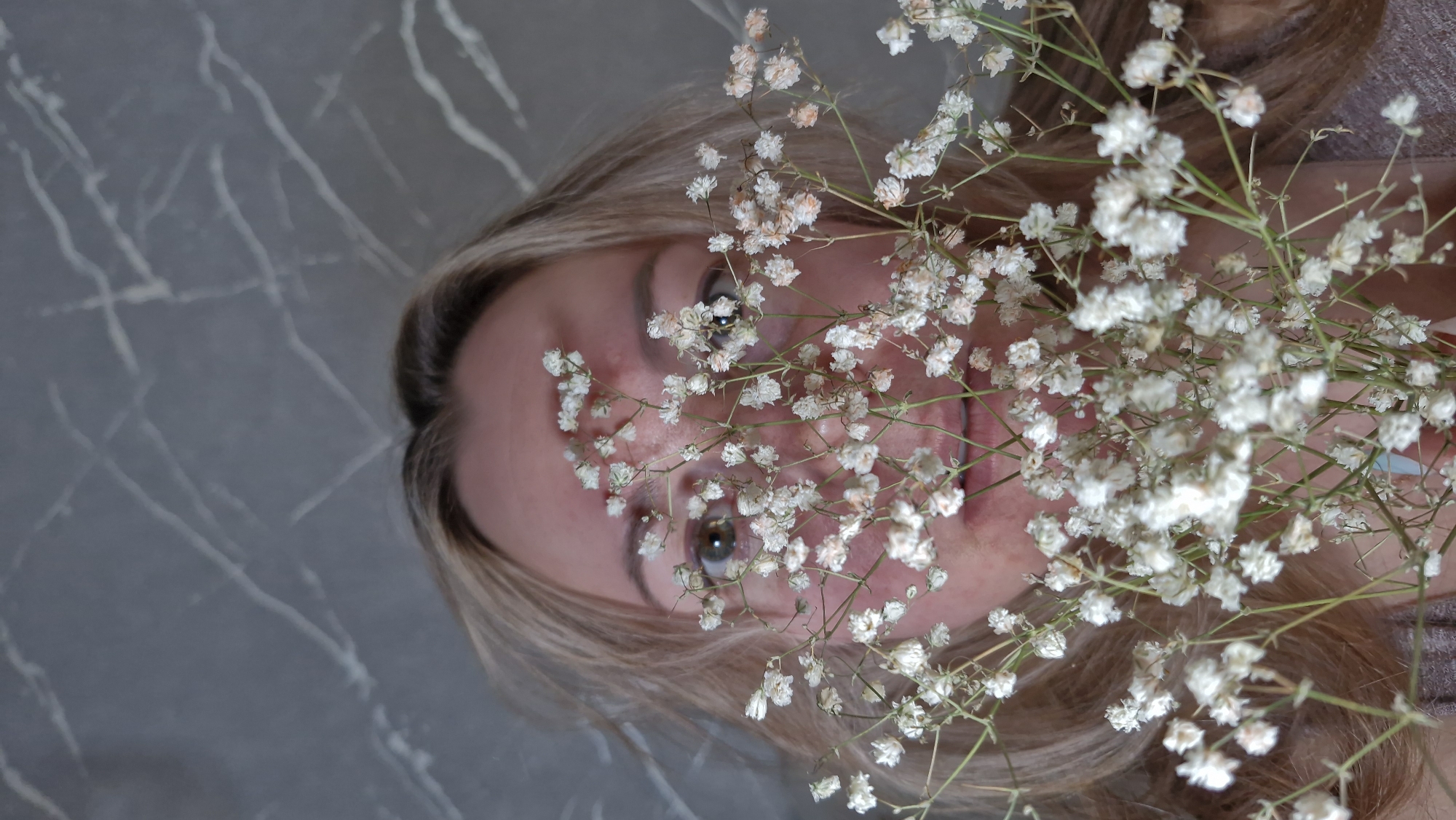 A woman is depicted in a close-up, top-down shot, lying on a dark gray surface with white, marble-like veining. Her head is positioned such that her gaze is directed upwards towards the viewer. Her light blonde or light brown hair frames her face. A generous cluster of delicate white baby's breath flowers partially covers her face, with petals and stems resting across her forehead, eyes, and cheeks. Her brown eyes are open, reflecting a soft light, and a subtle, serene smile can be seen on her lips, conveying a gentle expression. The focus is sharp on the woman's face and the flowers, while the background surface provides a sophisticated, textured setting. There is no discernible text. The image's context places its location in Otchor, Russia, although no visual cues within the photograph itself confirm this.