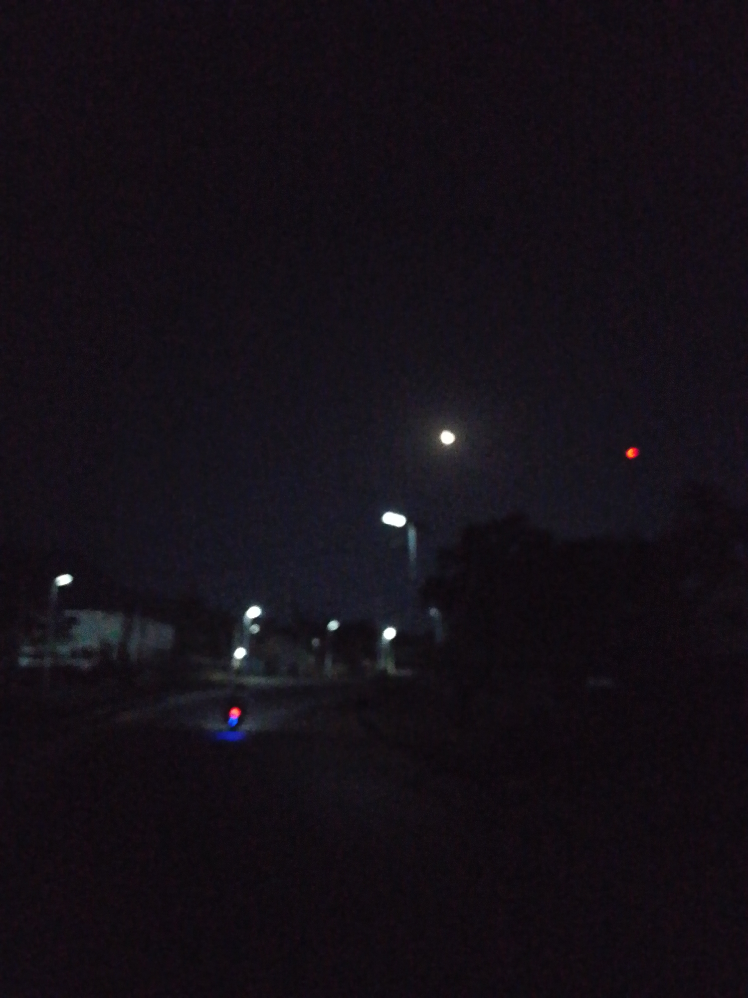 The image depicts a night scene in Adankolo, Nigeria. The environment is dark, with the light source appearing to be dim streetlights and a moon. A road stretches into the distance, and on the left is a building. There is a cluster of trees, and the red light could be on top of a building. At a closer distance, there seems to be a motorbike with the rear lights on. The image is blurred.