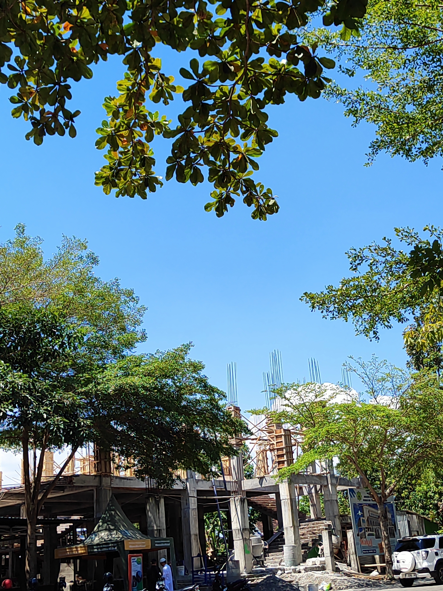 An eye-level, medium shot captures a construction site in Bondowoso, Indonesia, on a clear, sunny day. Lush green trees partially obscure the view, their leaves a vibrant contrast against the brilliant blue sky. In the foreground, leafy branches of a large tree overhang from the top of the frame. The partially constructed building features concrete pillars and beams, with rebar protruding from various levels. Wooden scaffolding is visible around some sections of the structure. In the mid-ground, a small, green, tent-like structure stands near the entrance of the building. A white SUV is parked to the right of the construction site. Several individuals are present, some dressed in traditional white attire, suggesting religious or cultural activities. Motorbikes are parked nearby, indicating local transportation. The scene is bustling with activity, implying ongoing construction and daily life in the city.