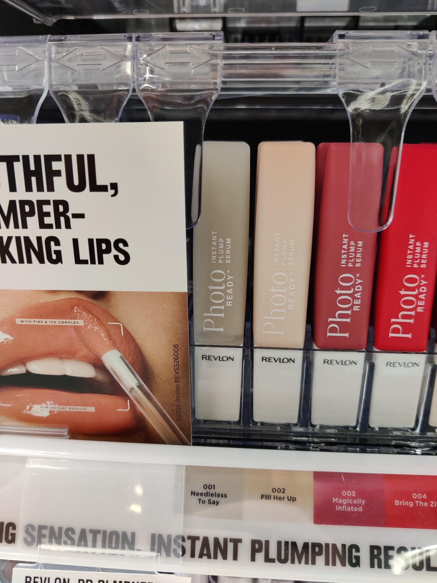The image shows a close-up, eye-level view of a retail display shelf featuring Revlon Photo Ready Instant Plump products. The display contains several lipstick-like tubes in various shades, including beige, pink, and red, arranged vertically behind clear plastic dividers. Each tube has "Photo READY™ PLUMP SERUM" printed vertically on it, and "REVLON" printed horizontally below. Below these tubes, a row of product name labels is visible, with the numbers 001, 002, 003, and 004 followed by descriptive names like "Needless To Say," "Fill Her Up," "Magically Inflated," and "Bring The Zi." A large sign partially visible in the foreground promotes "THFUL, MPPER- KING LIPS" and shows a close-up of lips with a gloss applicator. The overall environment appears to be a well-lit store aisle, suggesting a cosmetics section. The specific location of Mechanicsburg, United States of America, is not discernible from the visual information in the image.