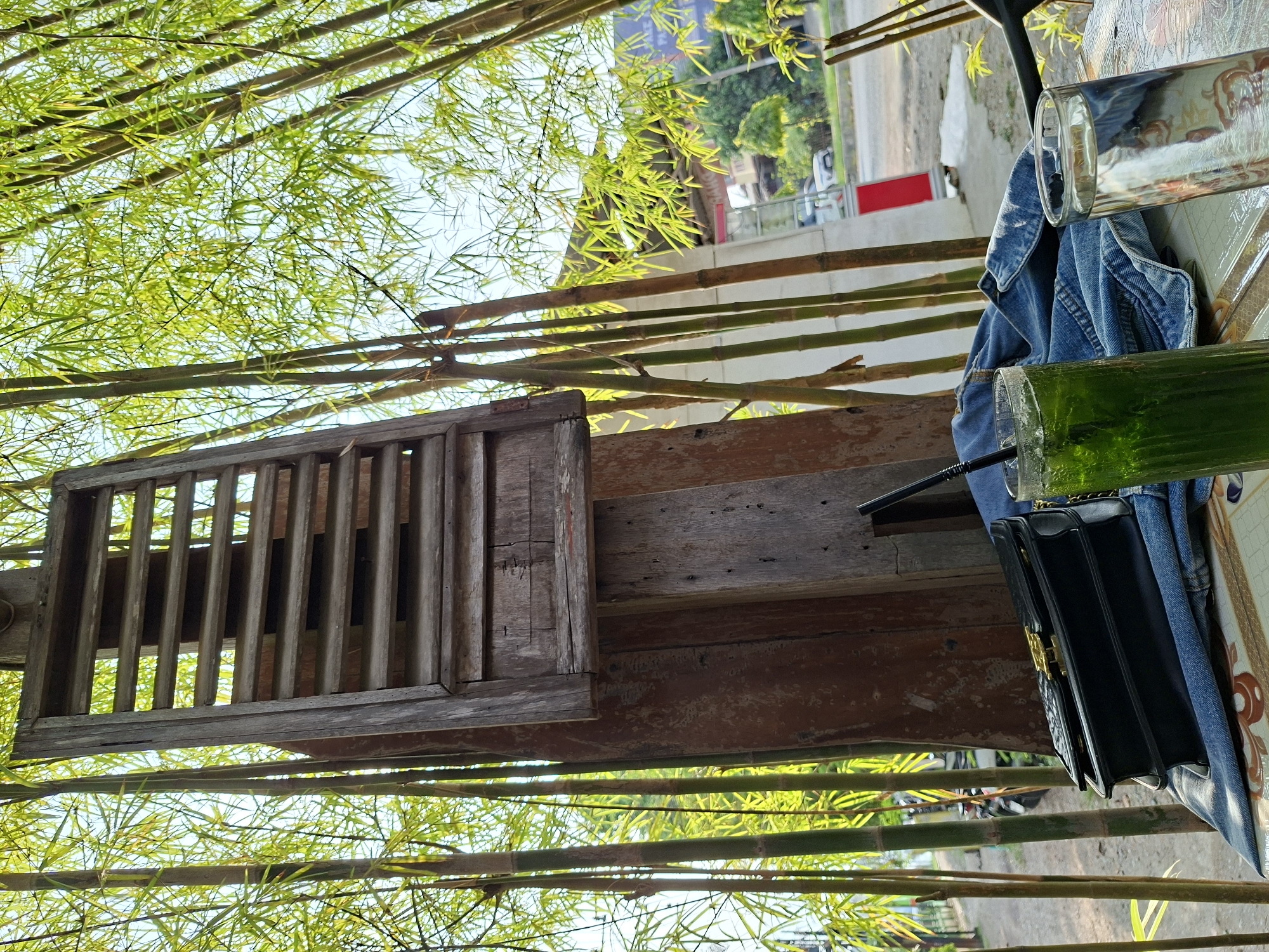 The scene is an outdoor café or restaurant located in Pantegajah, Indonesia. The perspective is from below, looking up at a structure made of bamboo and wood, likely a pergola or shaded area. Green bamboo stalks with lush leaves form the foreground and background, creating a sense of enclosure.
A wooden lattice window is attached to the structure, casting shadows on the wooden planks below. A table is partially visible in the upper right, with two glasses (one filled with a green liquid and a straw, the other empty) and a blue denim jacket. A black handbag is placed on the table. The overall setting suggests a relaxed and natural atmosphere.