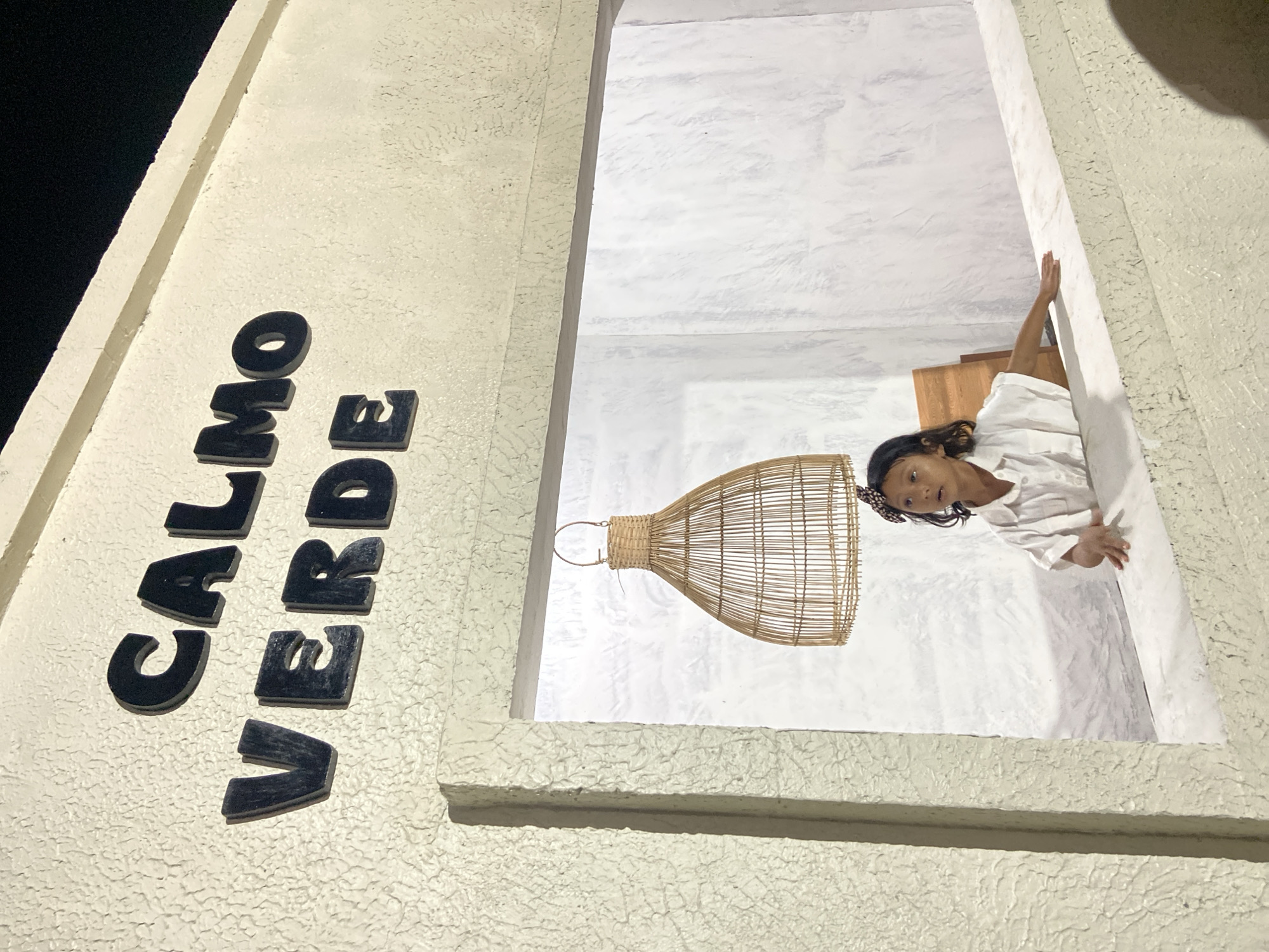 An outdoor shot at night shows a young girl peeking over a ledge at "Calmo Verde" in Kesiman, Indonesia. The girl, wearing a white shirt and a brown and white polka-dot hair bow, is looking towards the left of the frame. She is leaning on a white ledge in front of a textured white wall. Above her, a wicker lampshade hangs from the ceiling. The restaurant's name, "CALMO VERDE," is displayed in large black letters on the textured cream-colored exterior wall above the girl. The lighting suggests it is nighttime.