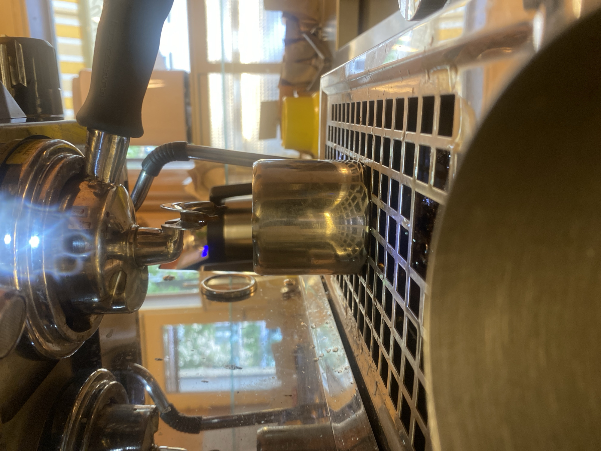The image shows a close-up, low-angle view of the drip tray area of a commercial espresso machine. A small, metallic shot glass is positioned on the metal grid of the drip tray, beneath the portafilter from which a stream of dark liquid is dispensing. The machine's chrome surface is reflective, showing glints of light and reflections of the surrounding environment, including windows with natural light and yellow awnings. The background is blurred but suggests an indoor setting, possibly a café. There are no people clearly visible in the frame, and the focus is on the coffee-making process. The scene is clean and functional, typical of a professional coffee preparation area. The location is not specified as Warsaw, Poland.