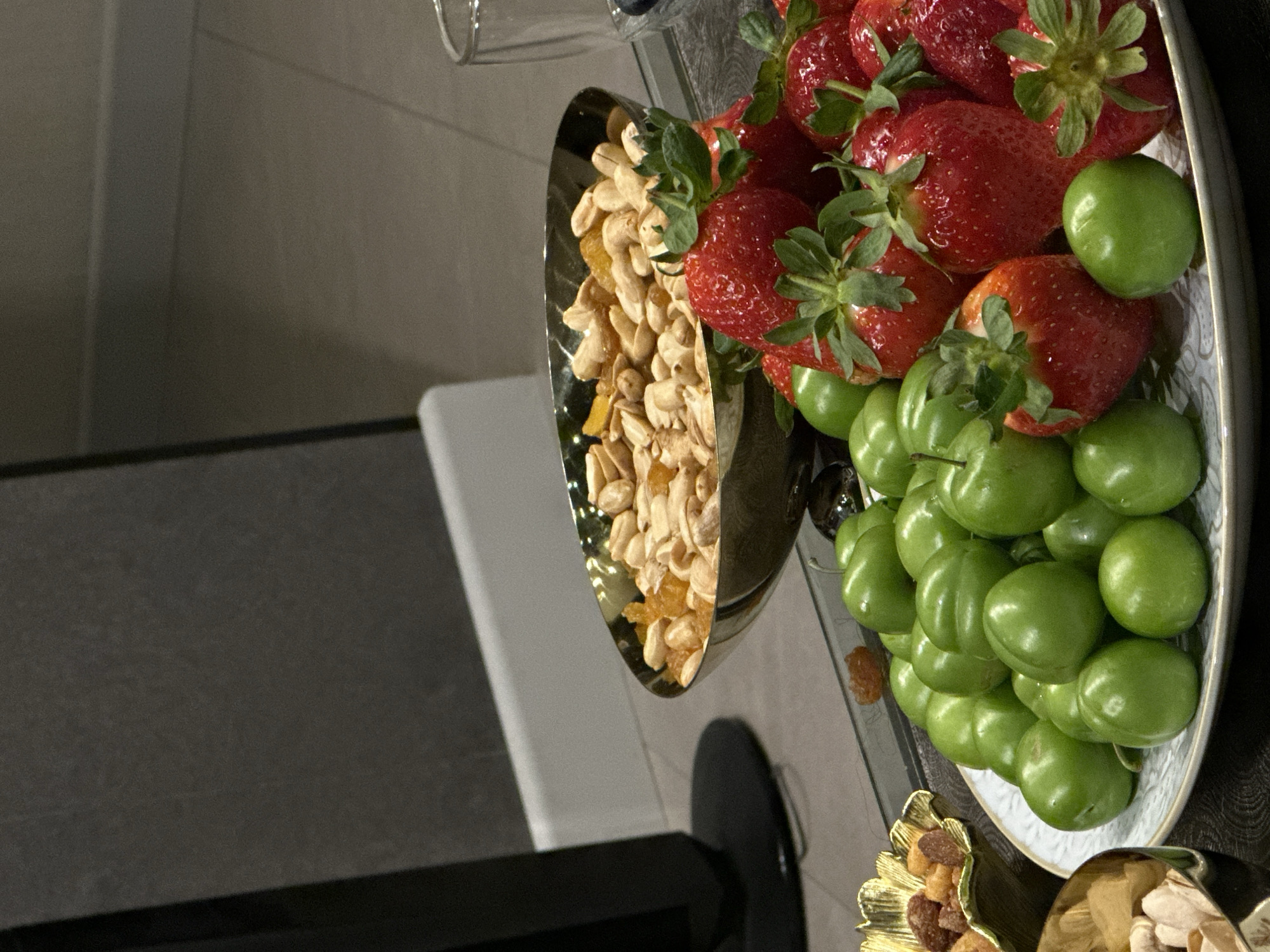 The image shows a silver platter with a variety of fruits and nuts. Strawberries, green plums, cashews, and some golden-colored dried fruits are visible. The platter appears to be on a reflective surface, possibly a glass tabletop. The background shows a light gray wall and a white chair or object. The location in the city of Yerevan, Armenia is not determinable from the image. There is no evidence of people, interactions or any explicit actions taking place.