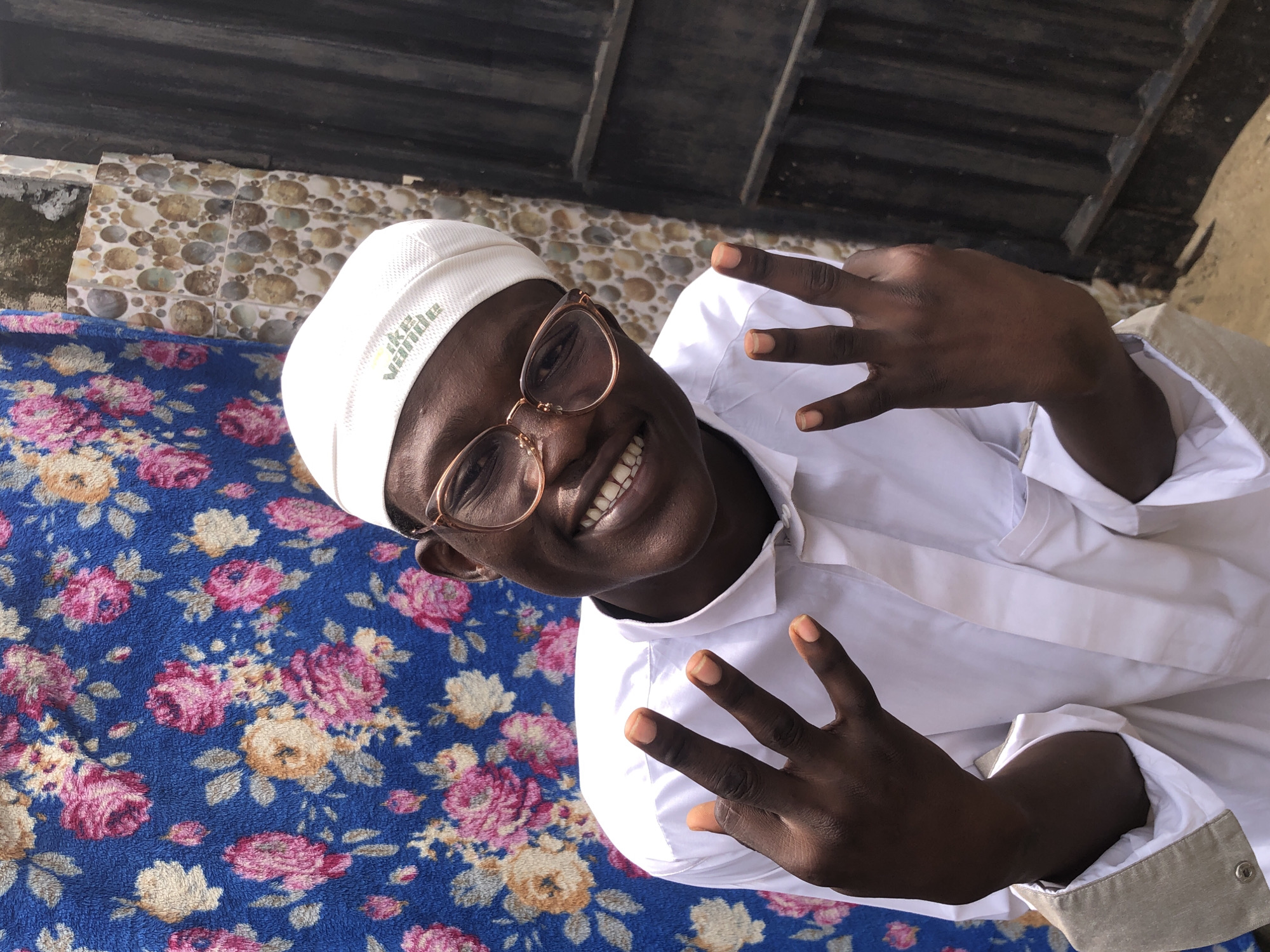 A young Black person wearing a white skullcap and glasses smiles at the camera. They are wearing a white collared shirt and have their hands raised, showing their fingers. The background is a blue floral fabric on the left and a wall with circular patterned tiles and a metal gate on the right. The scene appears to be outdoors in Ado, Nigeria, given the context.