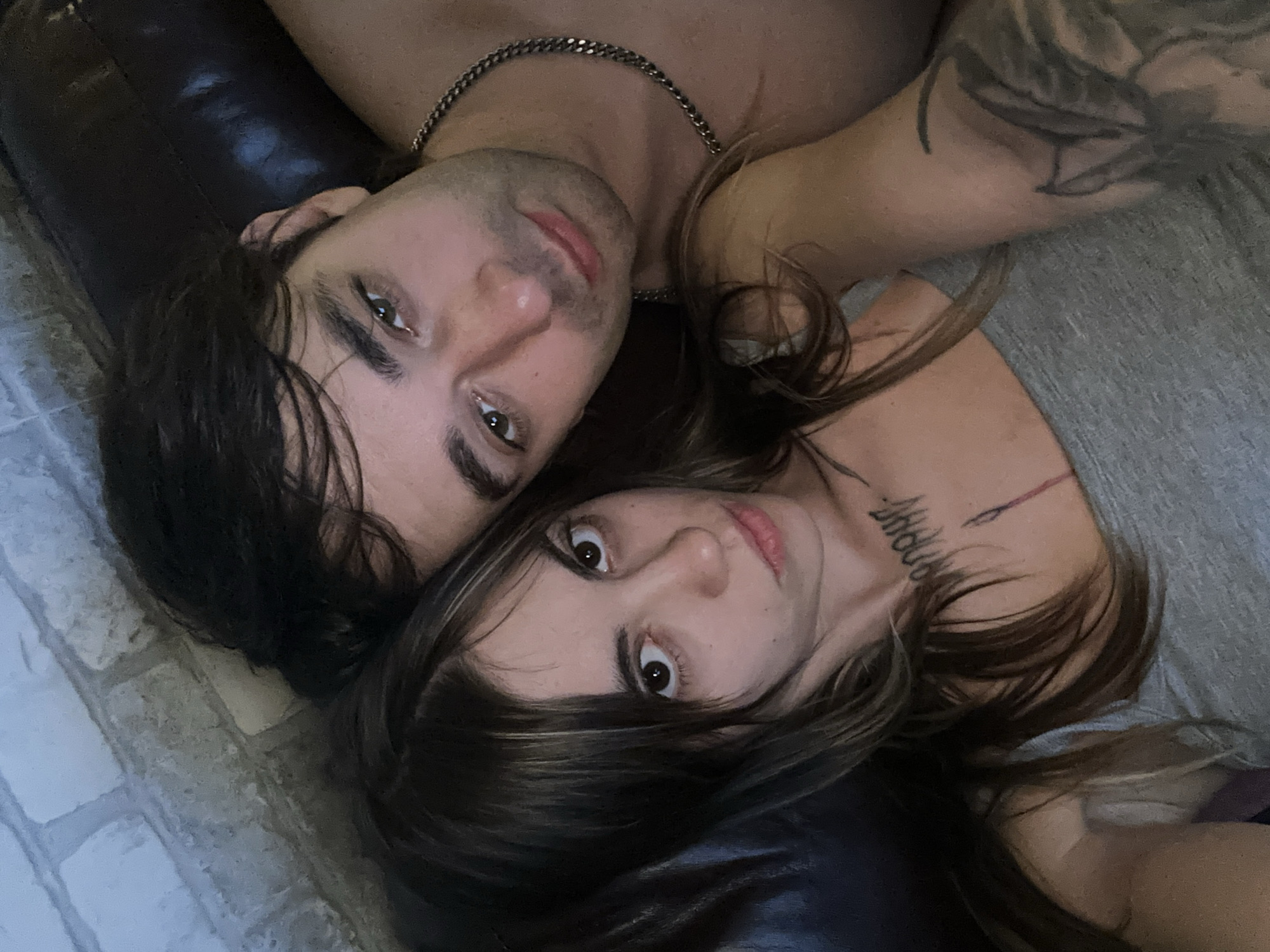 A close-up, high-angle selfie shows a couple lying down, their faces close together. The woman is on the left, facing the camera with her eyes open and mouth slightly parted. She has long, dark brown hair with blonde highlights and visible tattoos on her neck and arm. She is wearing a grey tank top. The man is to her right, also looking towards the camera. He has dark hair and is wearing a silver chain necklace. His eyes are open, and his lips are slightly pursed. They are positioned on what appears to be a dark leather couch, with a brick wall visible in the background. The lighting is dim, suggesting an indoor setting. There is no discernible information about the location being Moscow, Russia.