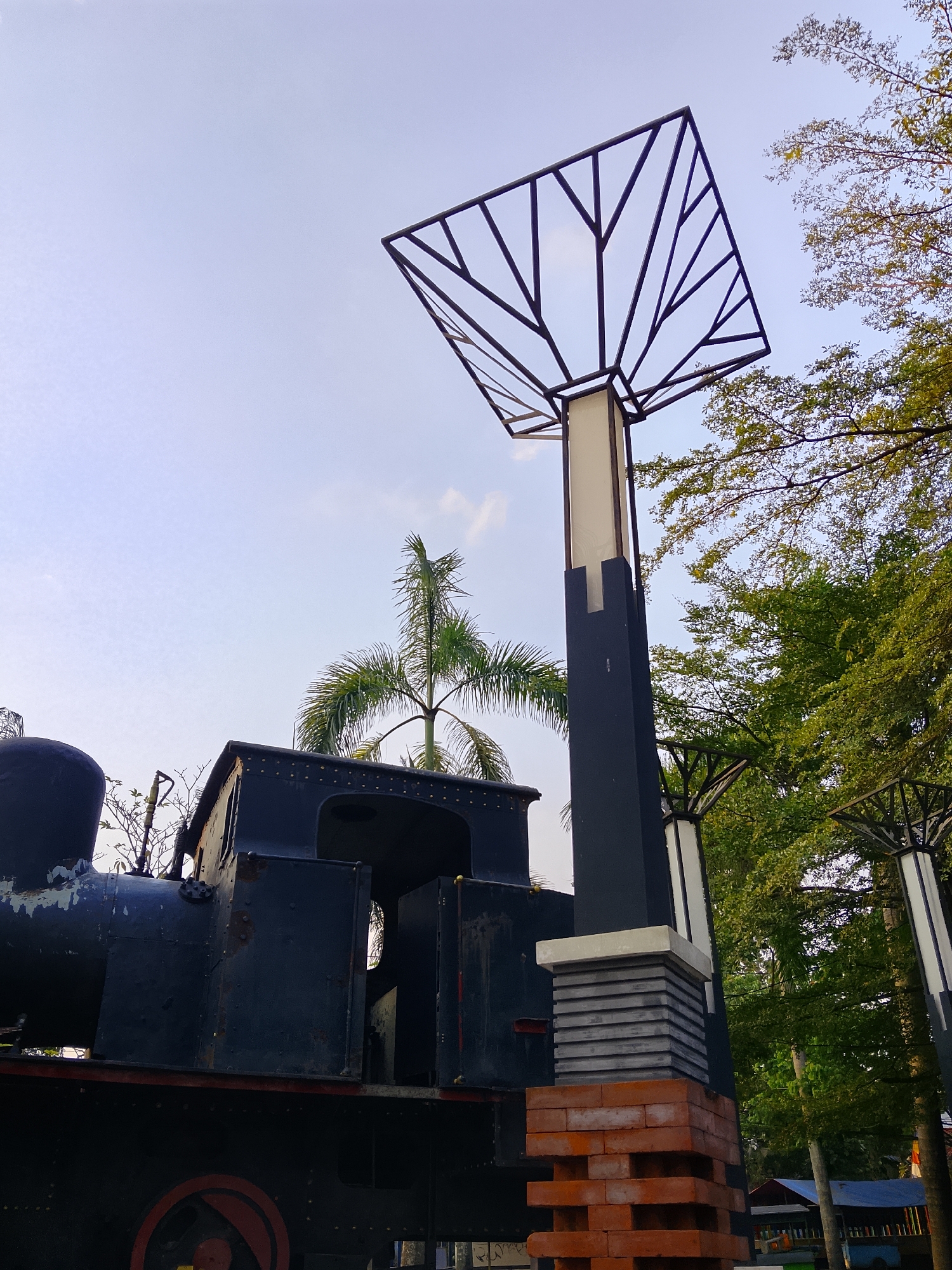 In Balun, Indonesia, an old, dark-colored train engine is parked in an outdoor setting. The train is positioned on the left side of the frame and takes up a significant portion of the lower half of the image. To the right of the train, a tall, modern lamppost with a black rectangular base and a white rectangular section stands prominently. The lamppost is topped with an abstract, geometric metal structure. Behind the lamppost and the train, lush green trees are visible, suggesting a park or garden environment. The sky above is a pale blue with a few wispy clouds. The overall scene depicts a blend of industrial heritage and modern design elements within a natural landscape.