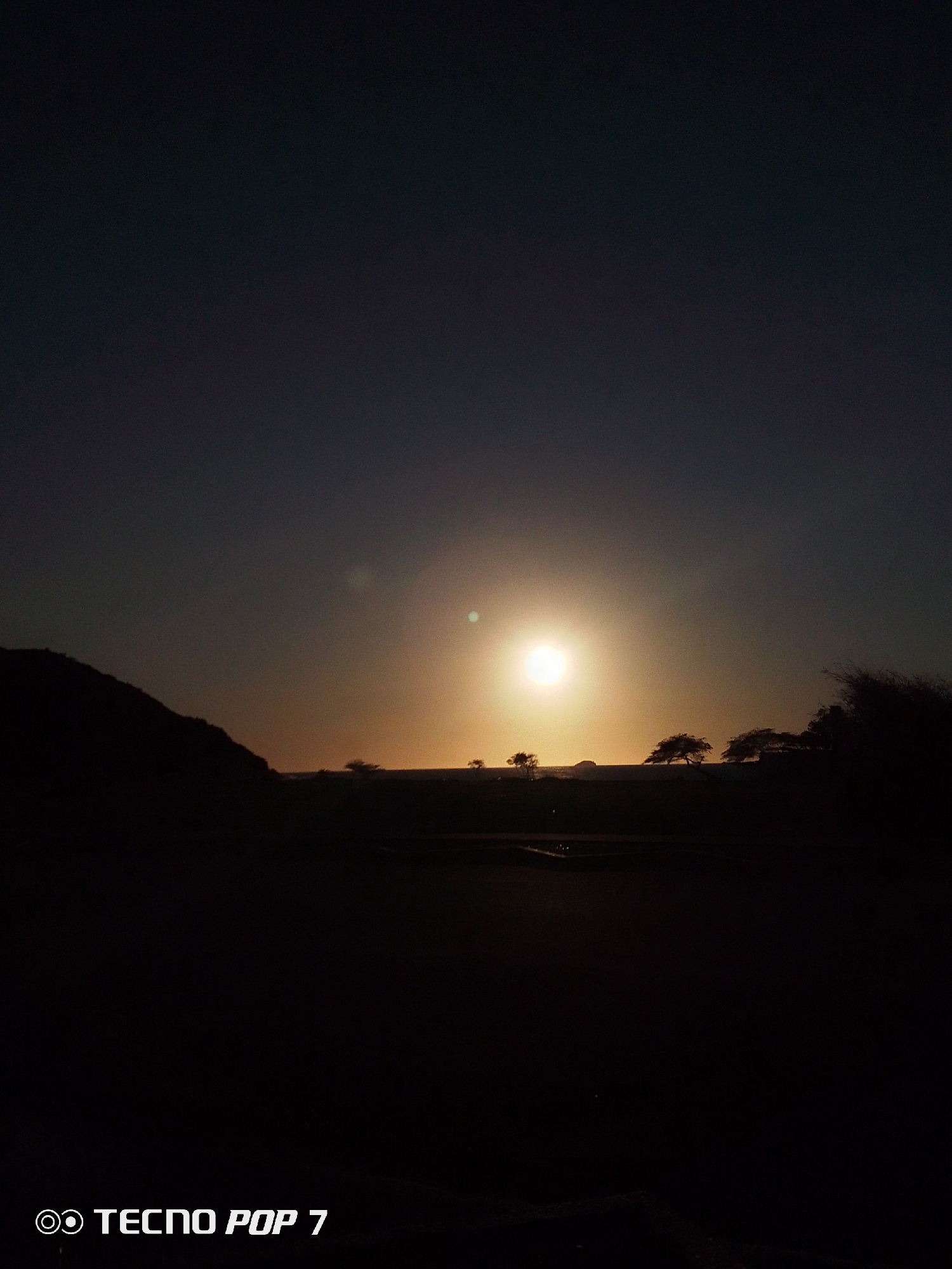 The image depicts a sunset scene in an unknown location. The sun is positioned in the center of the frame, casting a warm glow across the horizon. The foreground is dominated by dark silhouettes of land and vegetation, including a small hill on the left side and several trees scattered across the horizon. The sky transitions from a dark blue-gray at the top to a lighter orange-yellow near the sun. The bottom left corner contains the text "TECNO POP 7".