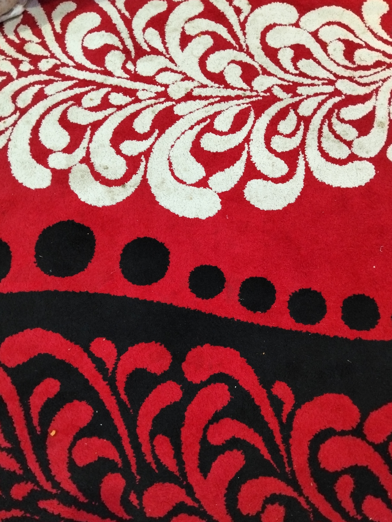 This is a close-up, overhead shot of a rug with a red background and a pattern. The pattern is divided into three horizontal sections. The top section features a swirling white design on red. The middle section is solid red with a row of black dots. The bottom section has a black background with a red swirling pattern. The rug appears to be indoors, possibly in a home. There are no people or other significant objects visible in the image. The location in the city of Cottrueng, Indonesia is not discernable from the image itself.