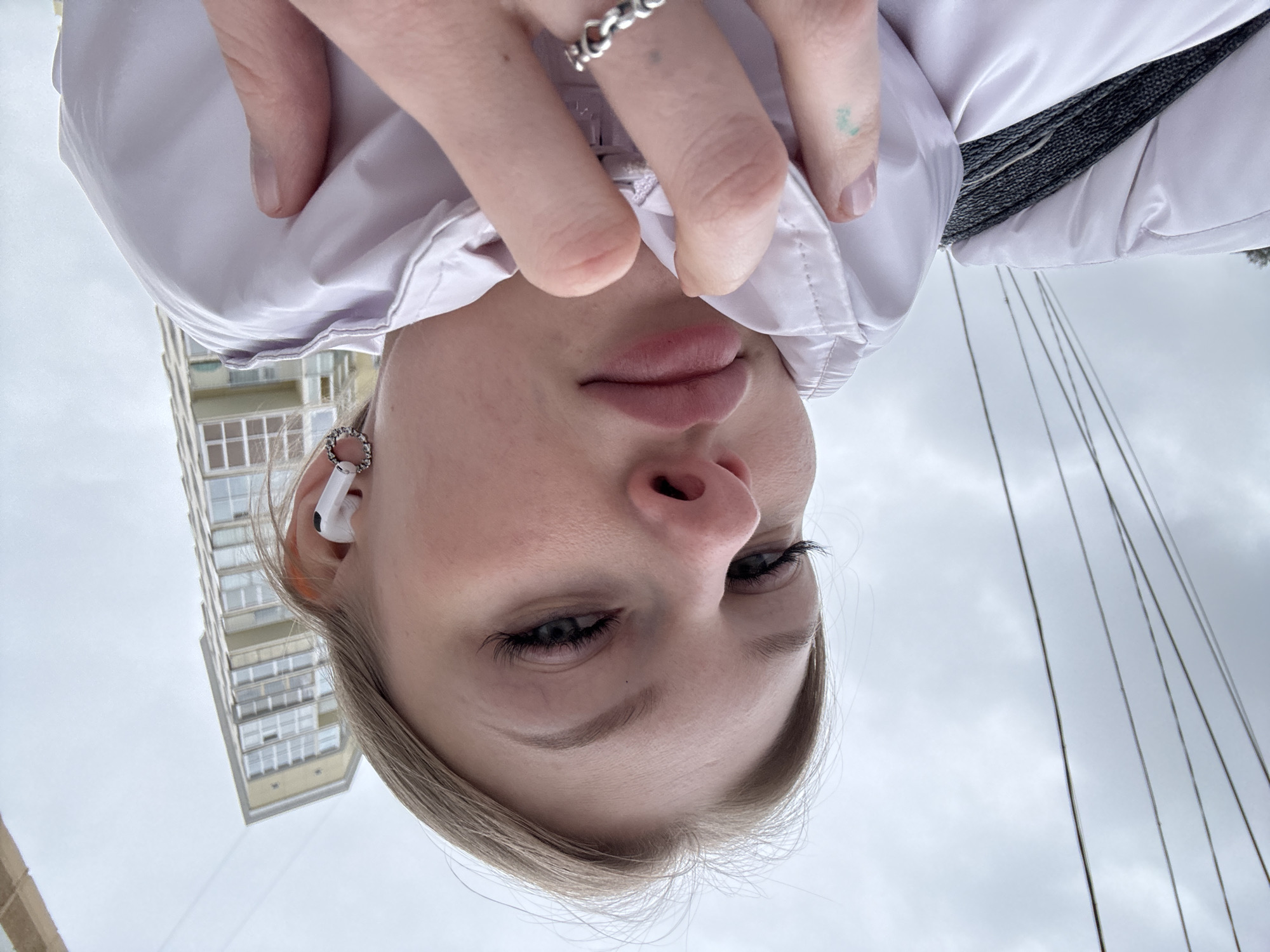 The image is an upside-down selfie of a young woman with light skin and blonde hair. She has an earbud in her left ear and a ring on one of her fingers. She is wearing a light-colored jacket. In the background, there is a building and what appear to be wires against a cloudy sky. Based on the architectural style of the building, the city is Perm, Russia. The woman appears to be holding up her jacket to partially cover her face.