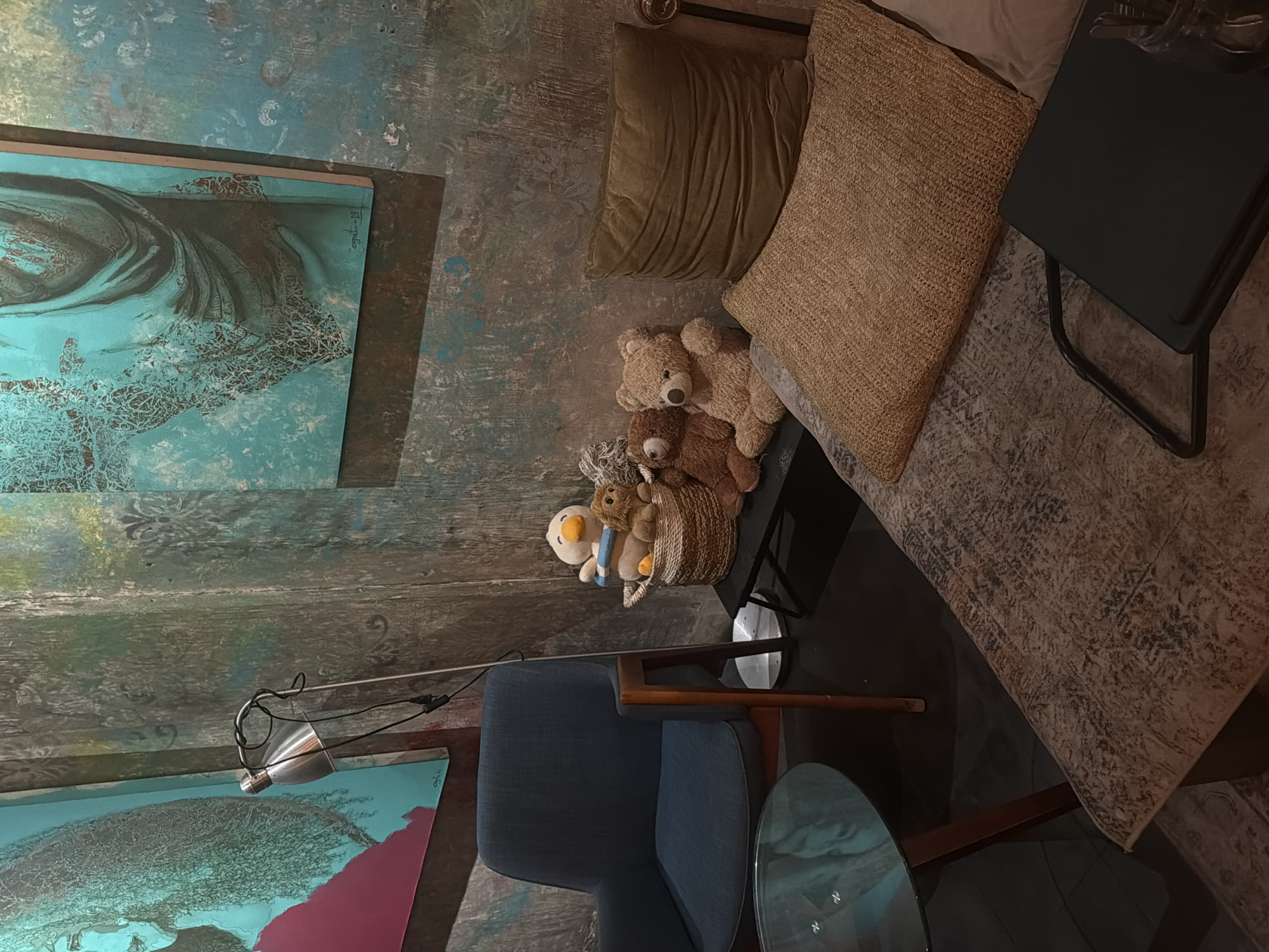 The image depicts a room with a bohemian aesthetic, likely in Ubud District, Indonesia, characterized by a textured, teal-colored wall adorned with two framed abstract paintings. A floor lamp casts light upon a blue upholstered chair positioned beside a small, glass-topped side table. Adjacent to the chair, a bed or seating area features several pillows in varying shades of brown and beige. A collection of stuffed animals, including teddy bears, is arranged near the edge of the bed or seating. A small black side table sits nearby. The overall atmosphere suggests a cozy and artistic space.