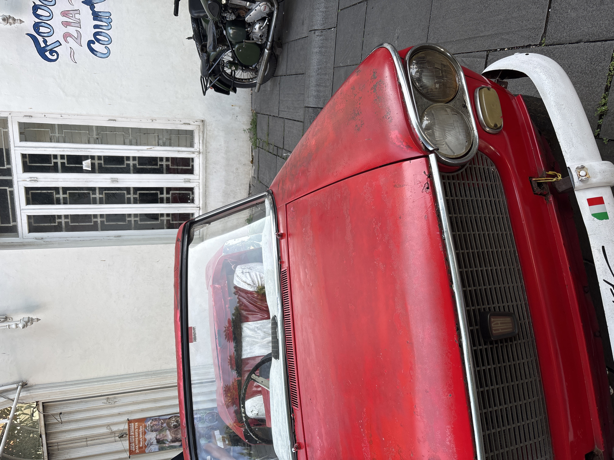 This image shows a red, vintage-style Fiat car parked on a paved surface, likely a street or sidewalk. A motorcycle is also parked in the background. The car is parked in front of a building with a white wall, a white window, and a sign that reads "Food 21A Court". On the left side of the picture, there is a window with a poster that says "JASA FOTO NYAAAA JASA FOTO KOTA TUA Hanya 5000/foto", indicating that it is in the Kota Tua (Old Town) area of West Jakarta, Indonesia. The car appears to be slightly weathered.
