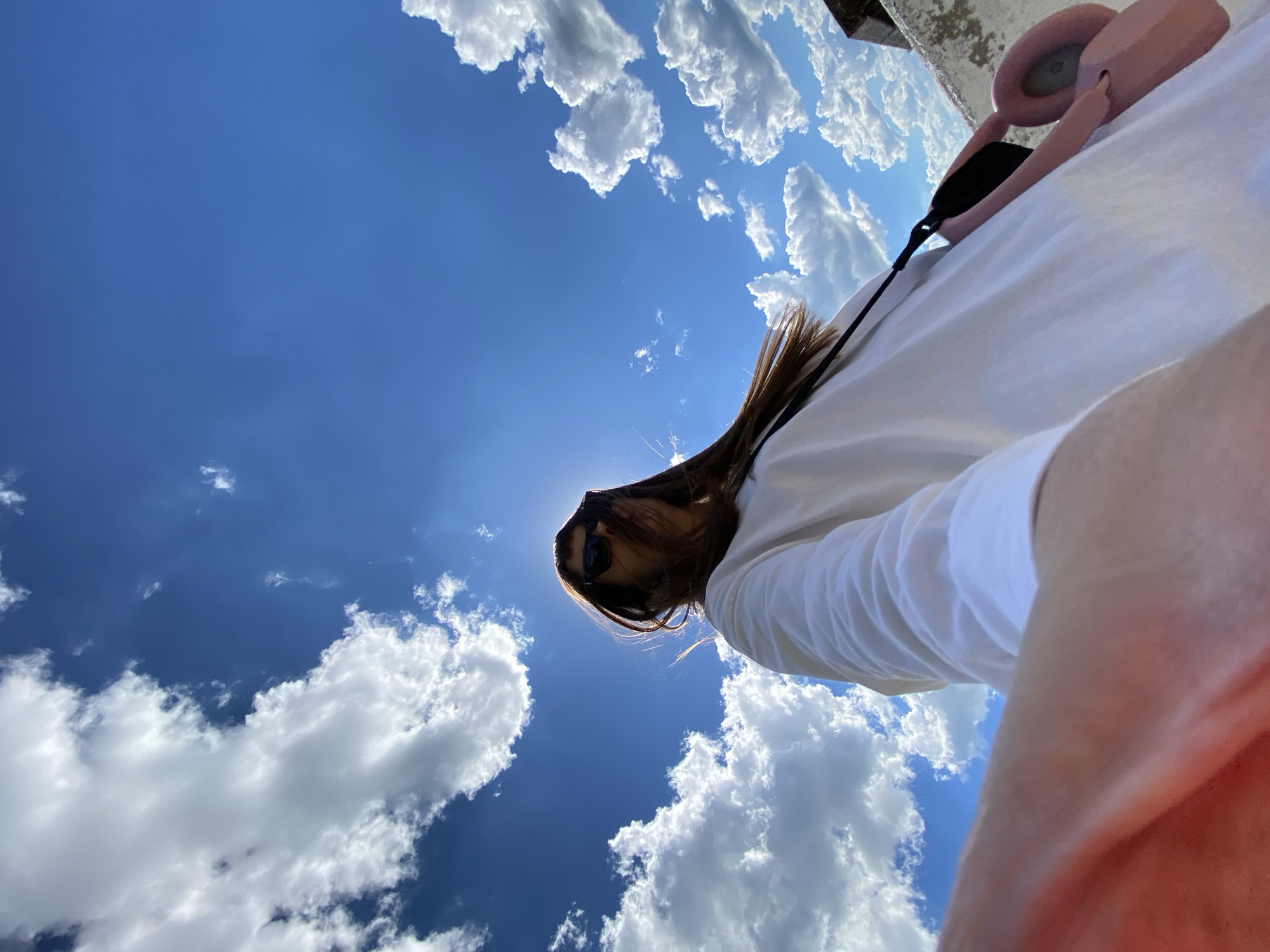 The image depicts a low-angle, wide shot of a person taking a selfie against a bright blue sky with scattered white clouds. The person, wearing a white long-sleeved shirt and sunglasses, is positioned slightly off-center, with their face mostly obscured by their long, brown hair and the glare of the sun. A pink bag with a black strap is visible over their shoulder. In the foreground, a portion of the person's arm or hand is visible, also slightly out of focus. The background shows the edge of a rooftop or ledge, appearing textured and weathered. The scene appears to be outdoors on a sunny day. The location is identified as Votkinsk, Russia, though there are no specific visual cues in the image to confirm this location.