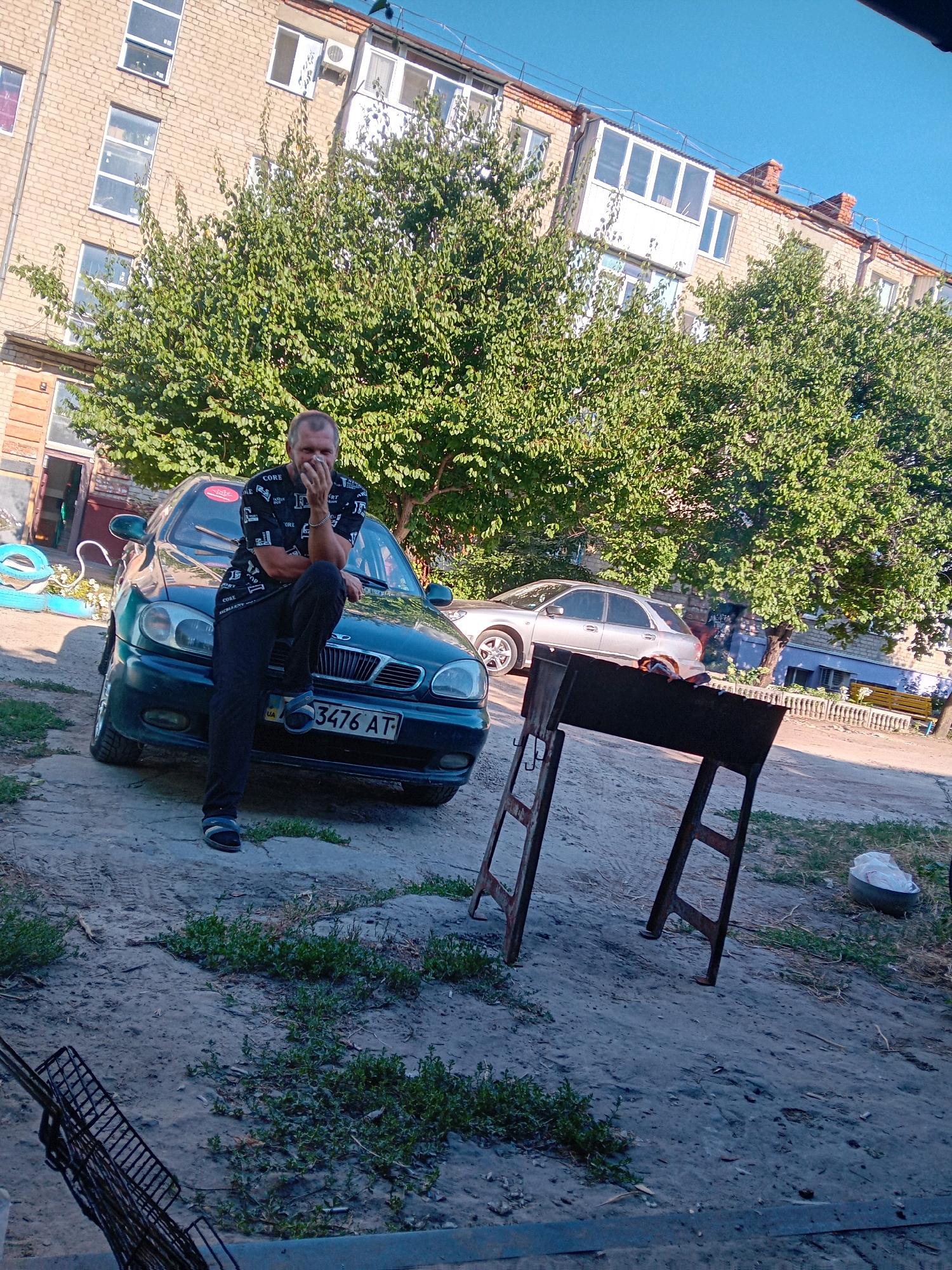 A man is leaning on a dark green sedan in front of a brick apartment building. He is wearing a black t-shirt with a repeating white pattern and black pants. He is covering his mouth with his hand. The car has a license plate that reads "3476 AT". A metal grill is set up on the dirt ground in front of the car. Trees with green leaves are visible behind the car and building. The sky is blue and clear. The scene appears to be an outdoor courtyard in the city of Chuhuiv, Ukraine.