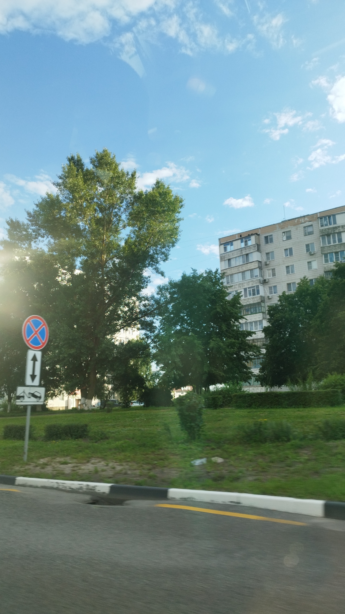 The image depicts a view from a vehicle on a sunny day, likely in a residential area of Stary Oskol, Russia. In the foreground, a road with a yellow line and a curb with alternating black and white markings is visible. To the left, there is a traffic sign with a red circle and a blue cross, indicating no stopping or parking, and below it, an arrow pointing up and down, suggesting traffic direction. Further left, another sign displays icons of a car being towed and a disabled vehicle. The middle ground features a grassy area with several trees, one large tree with lush green foliage prominently positioned on the left. In the background, a multi-story apartment building with numerous windows stands against a bright blue sky with scattered white clouds. The scene suggests a typical urban landscape with residential buildings and vegetation.