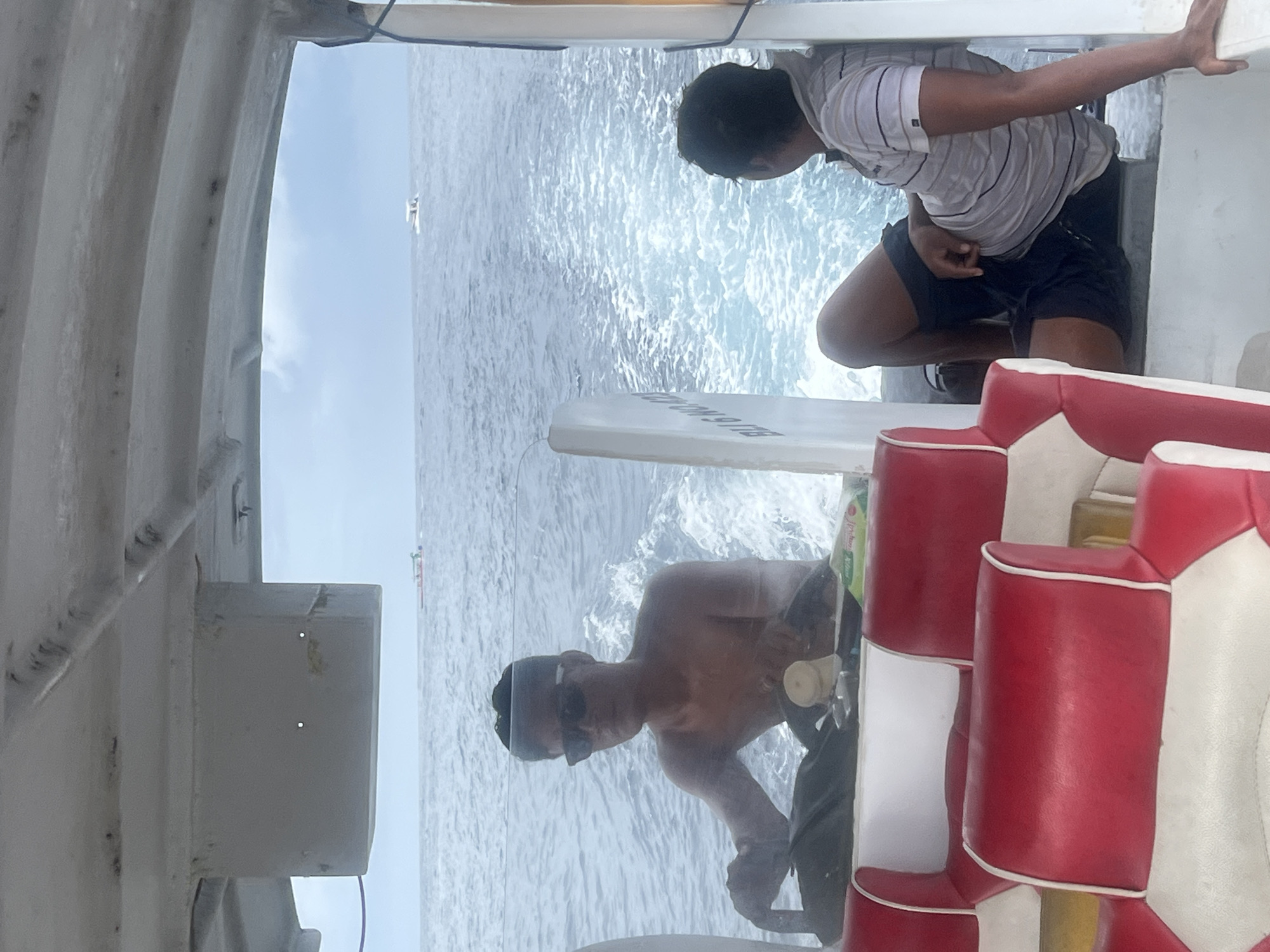 The image shows two shirtless men on a boat, likely taken on or near the waters of Nusa Penida, Indonesia. One man, wearing sunglasses, is closer to the camera, while another man, dressed in a striped shirt and dark shorts, is leaning over the edge of the boat looking at the water. The boat's wake is visible in the background, suggesting it's moving through the water. A portion of the boat's interior, including white and red cushioned seats, is visible. The overall environment is maritime, indicative of a coastal or island setting.