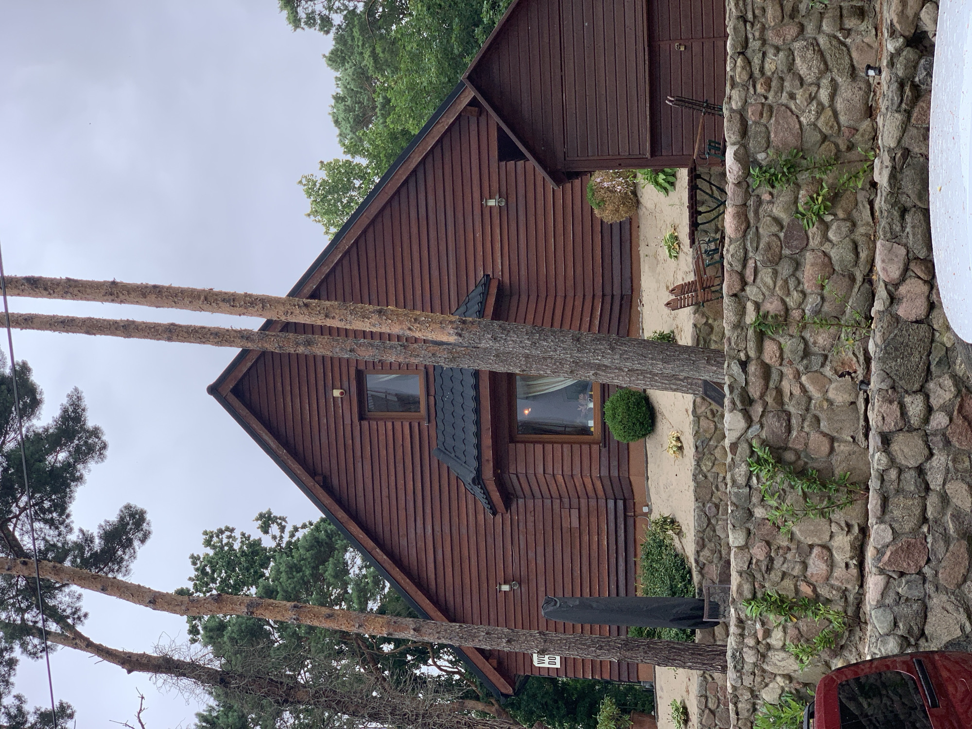 The image shows a wooden house in the city of Pobierowo, Poland. The house has a dark brown, horizontal wooden siding and a dark, tiled roof. There are two windows visible on the front of the house, one smaller upper window and a larger lower window. A dark-colored outdoor umbrella is leaning against the side of the house. In front of the house, there is a low, multi-layered stone wall. Behind the wall, there is a small outdoor seating area with a table and two chairs. There are several tall pine trees in the foreground and background, as well as lush green foliage. The sky is overcast. A red vehicle is partially visible in the lower left corner of the image.