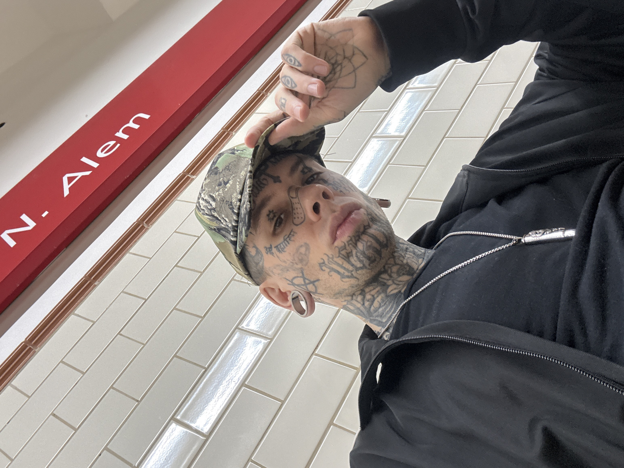 The image is a low-angle selfie of a tattooed individual wearing a camouflage baseball cap, a black zip-up hoodie, and a silver chain. The person has facial tattoos and stretched earlobes. Their left hand is raised, touching the brim of the cap, revealing additional hand tattoos. The background features white subway tiles and a red sign with white lettering that reads "N. Alem", which indicates the location is the Leandro N. Alem subway station in Buenos Aires, Argentina.