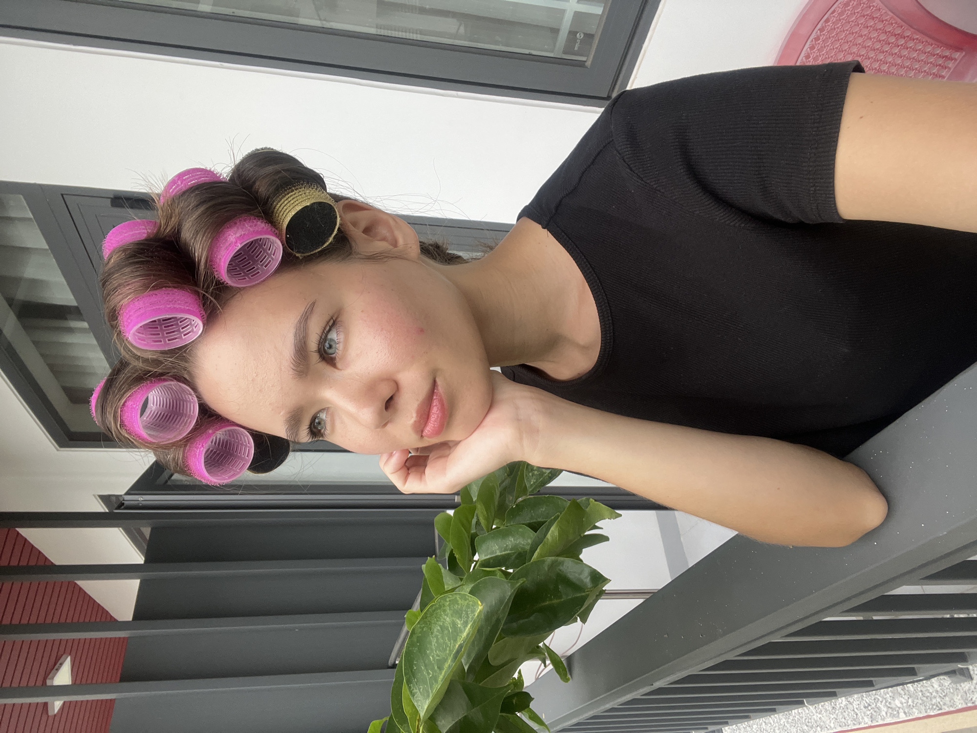 The image depicts a young woman with fair skin and blue eyes, her hair styled in rollers, some pink and some gold, wearing a black t-shirt. She is resting her chin on her hand, leaning on a gray metal railing, with a potted green plant in front of her. The background includes a white wall, a window, and glimpses of a red and gray structure behind the railing. Based on the prompt, the location is in Nam Nha Trang Ward, Vietnam.