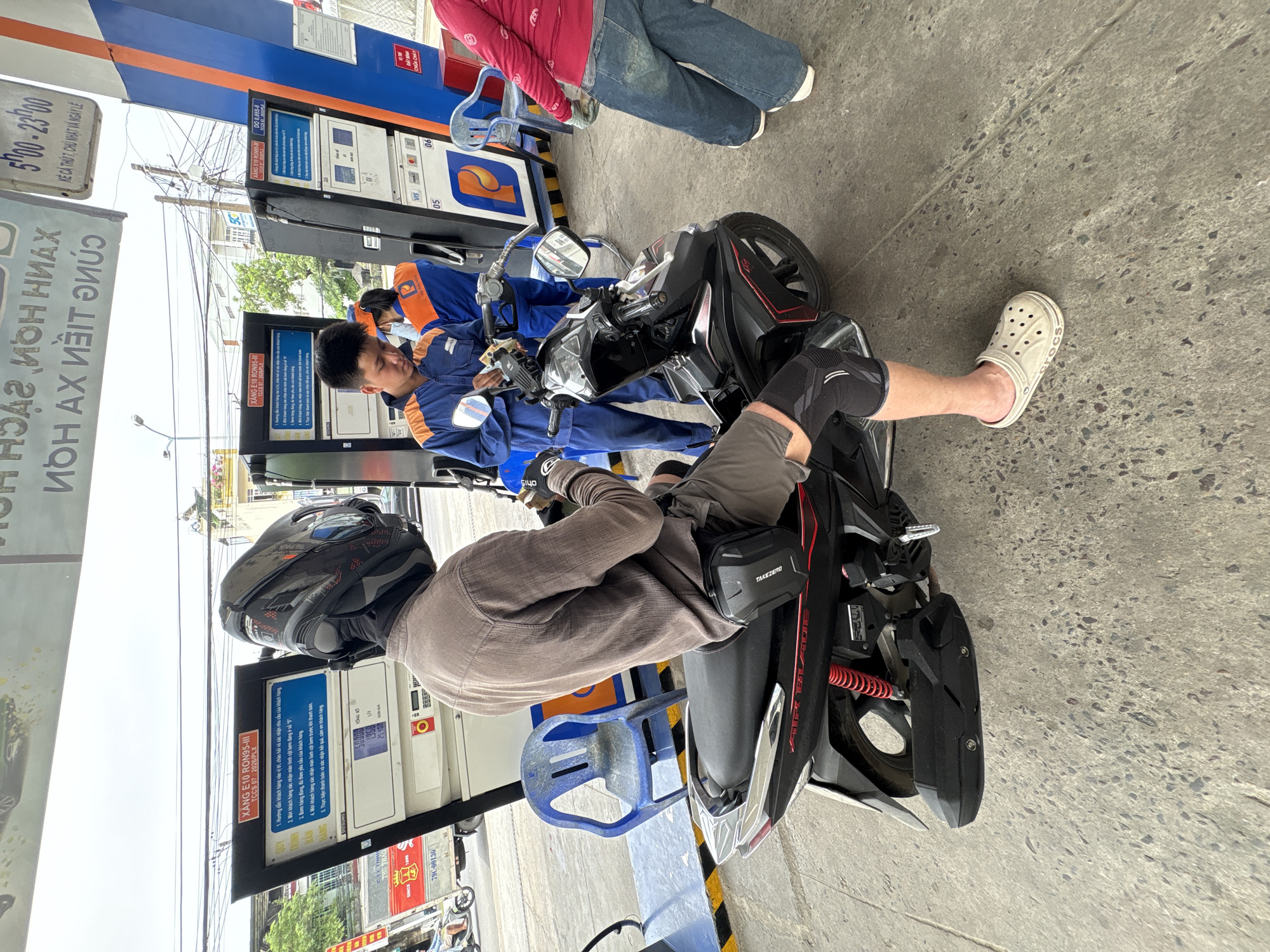 Here is a matter-of-fact description of the image:
The image shows a gas station scene, likely in Bac Nha Trang Ward, Vietnam, due to the text on the pump signage and its appearance. A motorcyclist wearing a helmet, knee pad, tan shirt, and crocs is seated on their bike. The motorcycle, a black Air Blade, is positioned next to a gas pump. A gas station attendant in a blue uniform is holding money and appears to be assisting another person on a scooter who is obscured by other pump equipment. Additional gas pumps are visible in the background. A blue plastic chair sits near the motorcycle. The environment shows overhead electrical wires and parts of surrounding buildings.