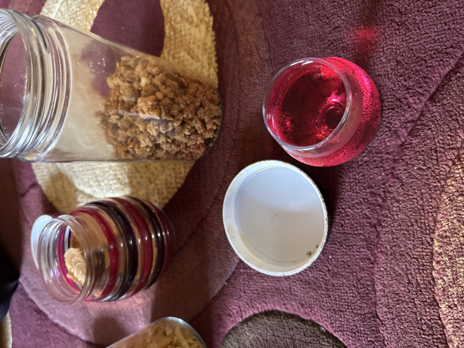 This image depicts a static scene within the city of Pante, Indonesia, showcasing several food-related items arranged on a patterned rug. In the foreground, a clear glass contains a vibrant red, effervescent liquid, possibly a beverage, showing small bubbles. Adjacent to it, a white, circular metal lid, likely from a jar, rests upside down on the carpet, exhibiting minor discoloration along its edge. Towards the mid-ground left, a small, clear glass jar with horizontal maroon and dark purple stripes contains a single, light brown, irregularly shaped snack, with its white plastic lid resting slightly askew on the rim. To the right of this, a taller, clear glass jar holds a quantity of small, light brown, granular or crumbly food items, filling approximately one-third of its volume. A portion of another clear glass jar, containing light-colored, thin snack items, is visible on the far left. The items are situated on a textured carpet featuring an abstract pattern of curvilinear and angular shapes in shades of deep red/maroon and beige. No individuals are present in the frame.