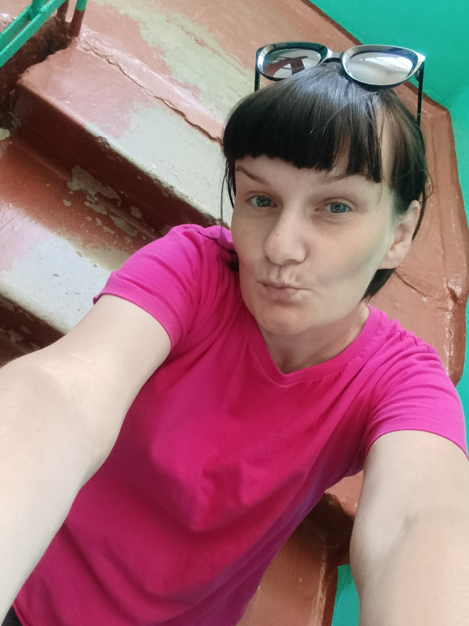 A woman in a pink t-shirt takes a selfie in a stairwell, likely within the city of Фоки, Russia. She has short, dark hair with bangs and is wearing sunglasses on her head. She is puckering her lips in a kissing face expression. The stairwell has brown and white painted steps, and the walls are partly painted in a bright turquoise color. The paint on the steps appears worn.