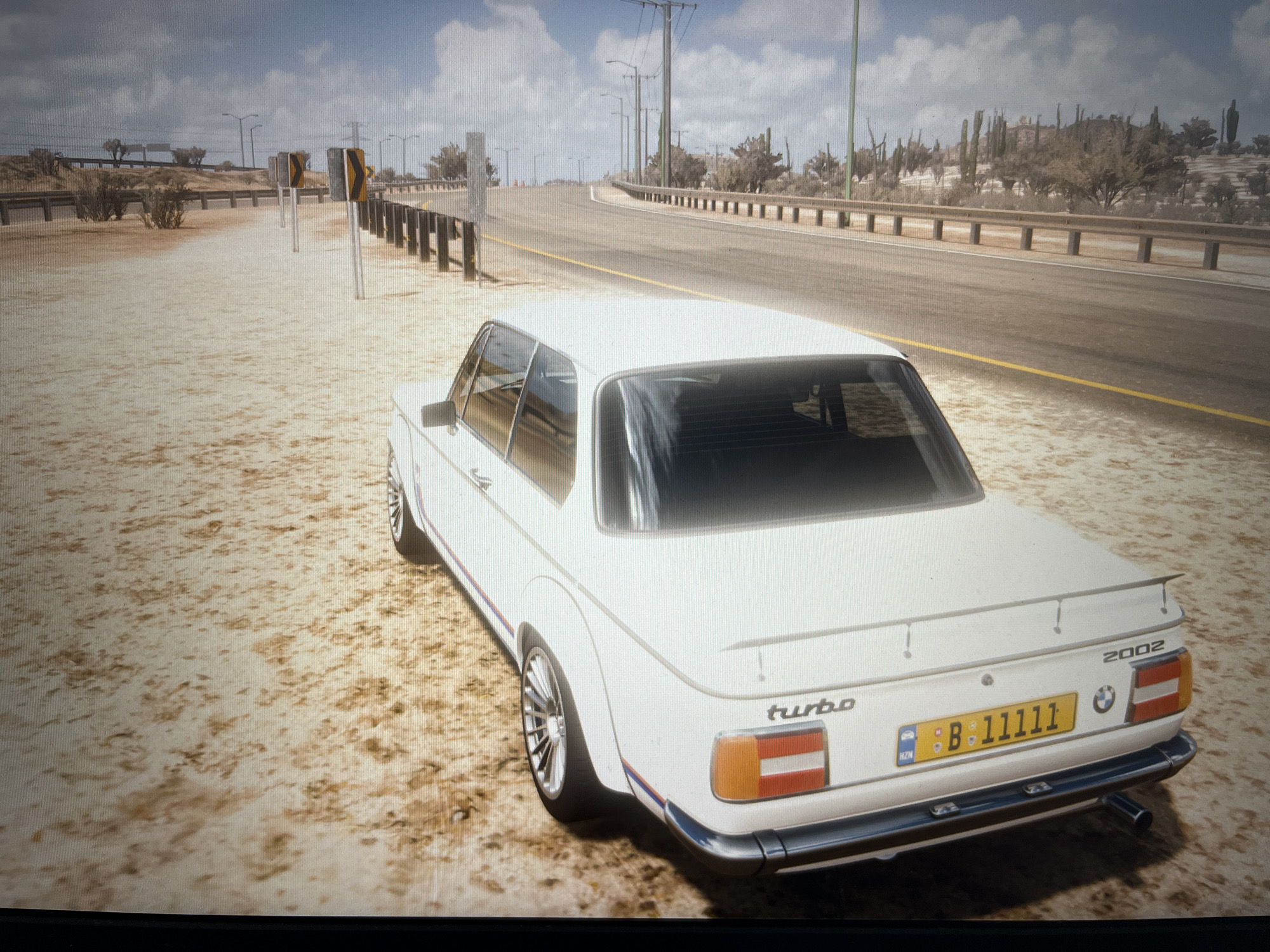 A white BMW 2002 Turbo is parked on a dirt shoulder next to a highway. The car is facing away from the camera, with its rear end visible. The license plate reads "B 11111". The highway is paved and has a yellow dividing line. The surrounding environment is dry and arid, with sparse vegetation and a dusty landscape. The sky is cloudy. The image is taken from a slightly elevated perspective, looking down at the car and the road. There are no people visible in the image. The scene appears to be a digital rendering, likely from a video game, given the slightly artificial look and the pristine condition of the car and road. There is no indication of the location being the city of Varna, Bulgaria; the arid landscape suggests a desert or semi-desert environment.