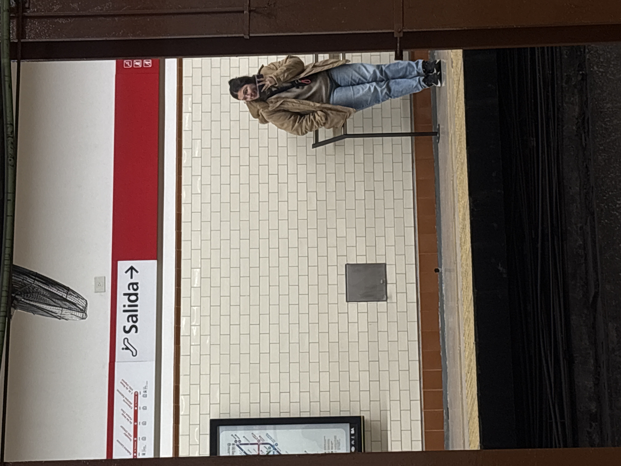 A person wearing a tan jacket and blue jeans is sitting on a bench at the Plaza de Mayo station in the Buenos Aires Subway (Subte). The station has white tiled walls with a red stripe and a "Salida" (Exit) sign. There is also a subway map, a fan, and a metal access panel on the wall. The photograph appears to have been taken from across the platform.