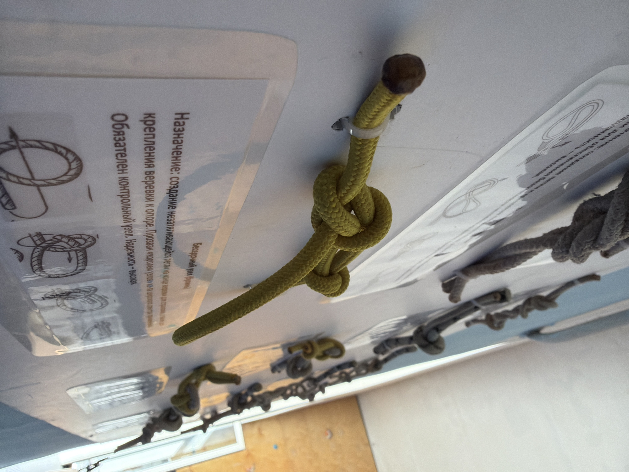 This is a close-up shot of a white wall displaying instructions and demonstrations of various rope knots. A yellow rope is prominently featured, tied in a knot and affixed to the wall with a plastic tie. Adjacent to the rope are printed instructions in Russian, providing details about the knot's purpose and importance. The scene also includes other knotted ropes and related instructional materials mounted on the wall. Based on the Russian text present in the image, it's likely the photo was taken in Russia. Based on other information, it is presumed to be taken in the city of Izhevsk.