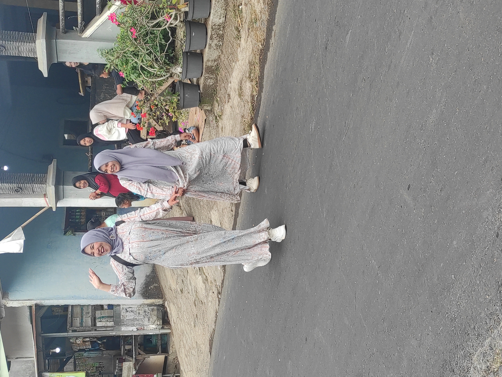 Two women wearing patterned dresses and headscarves walk hand-in-hand on an asphalt road. The woman on the left waves at the camera with her right hand and smiles broadly. The woman on the right also smiles. They are both wearing white sneakers. In the background, several people are gathered near a building with a light blue exterior and a porch. Some people are sitting on a bench, and others are standing. There are potted plants with pink flowers in front of the building. To the left of the road, there is a small shop with shelves stocked with items. The overall scene appears to be a residential or commercial street.