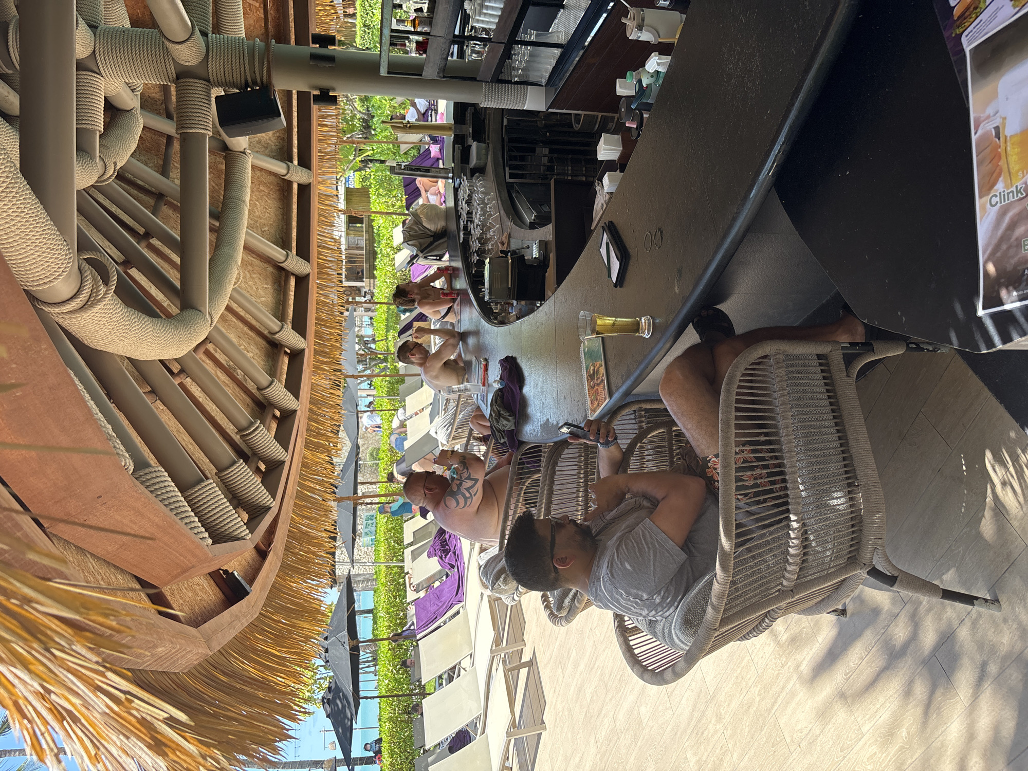 The image captures a bar setting in Baan Bo Phut, Thailand, where several patrons are seated along a dark-colored bar counter. The seating consists of woven chairs with visible details in the material. In the foreground, a man wearing glasses and a gray shirt sits in one of the chairs. His posture suggests he might be looking at or using a mobile phone. At the bar, drinks and other personal items are placed. Further down the counter, other people are sitting and engaging in various activities. Outside the bar, one can see a pool and beach area with chairs and umbrellas. The bar area is shaded by a thatch roof structure.