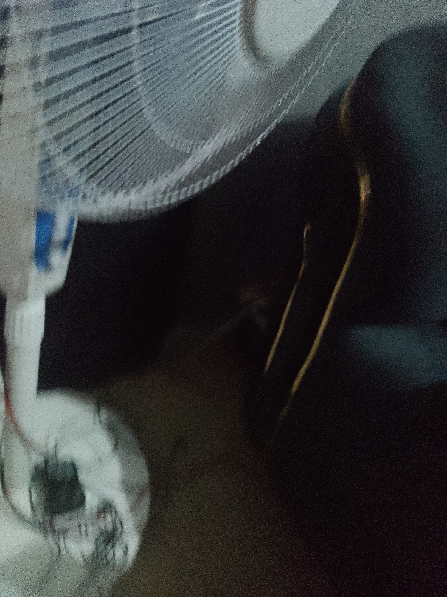 An extreme close-up, dimly lit, grainy shot focuses on a portion of an electric fan. The fan's grille, composed of many thin, white, parallel lines, occupies the upper left quadrant, angled away from the viewer. To the left, a white, cylindrical base of the fan is visible, with a blue accent. The lower portion of the base shows exposed wiring and electronic components. The right side of the image is dark, with a suggestion of a curved, dark object, possibly an arm or shoulder, illuminated by a faint, yellowish light that creates a highlight along its edge. The background is largely obscured by shadow. The scene appears to be indoors, given the presence of the fan and the artificial lighting.