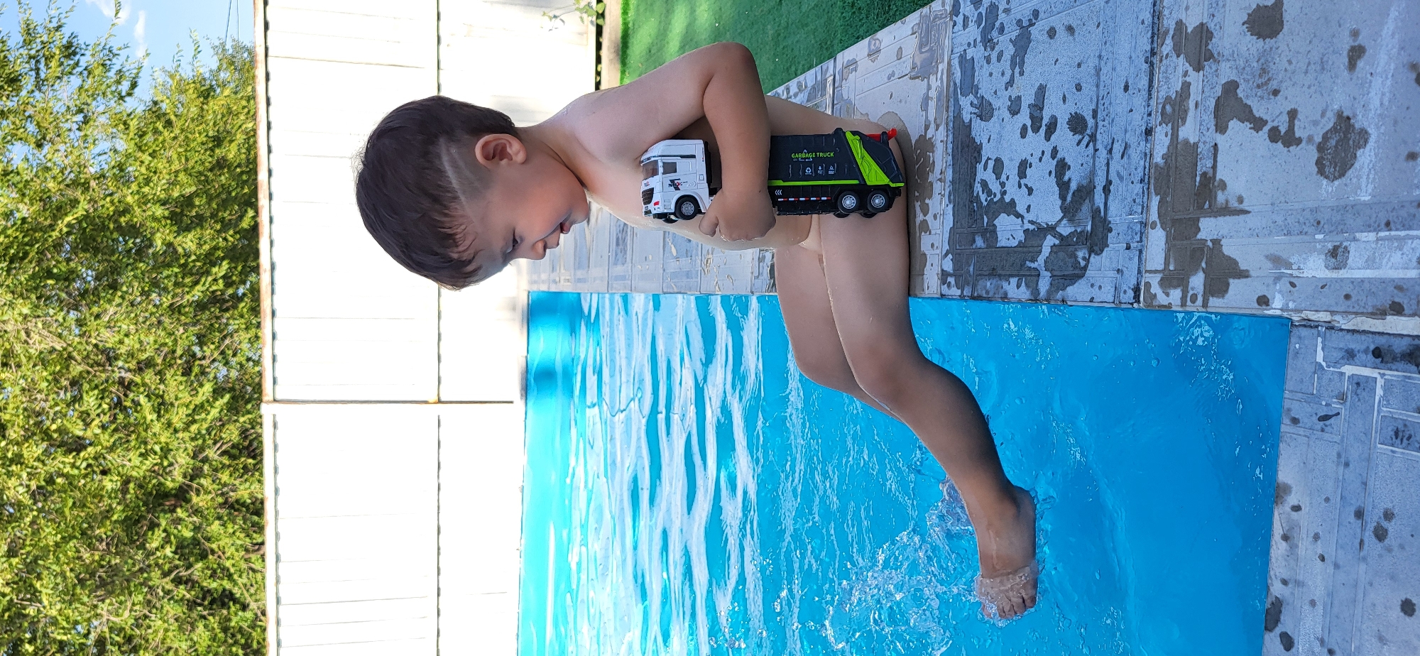 A young boy sits on the edge of a swimming pool with his legs dangling in the water, splashing it with his feet. He holds two toy trucks, one white and one black and green, in his arms. The background shows a white fence, green trees, and a blue sky. The boy has dark hair with a shaved design on the side. The scene appears to be a private backyard in Taldykorgan, Kazakhstan.
