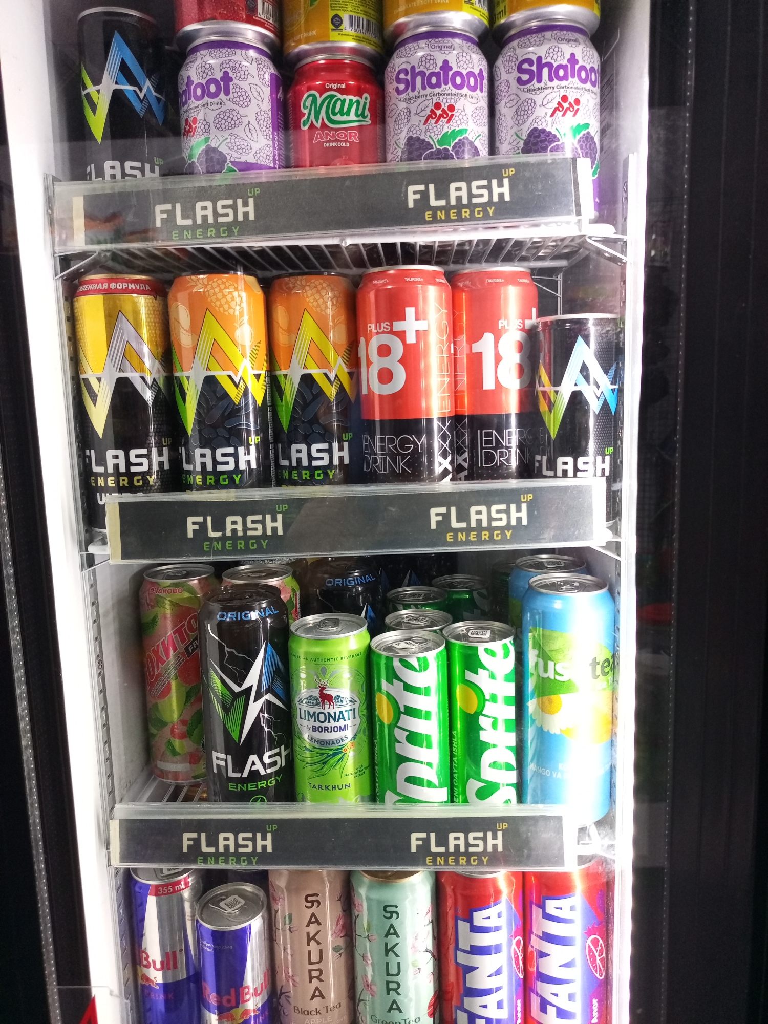 The image is a close-up, eye-level, interior shot of a refrigerator displaying an assortment of canned beverages. The refrigerator appears to be in a commercial setting, possibly a convenience store or a small market, located in the city of Kitab, Uzbekistan. The focus is on the shelves filled with various brands of energy drinks, sodas, and teas, with a prominent "FLASH ENERGY" branding visible on a divider. The cans are arranged neatly, with some brands appearing multiple times. Notable brands include Flash Energy, Shatoot, Mani, Plus 18, Fanta, Red Bull, Sakura, and Sprite. The lighting is bright and artificial, illuminating the products clearly. There are no people or actions depicted in the image. The environment is clean and organized, characteristic of a retail display.